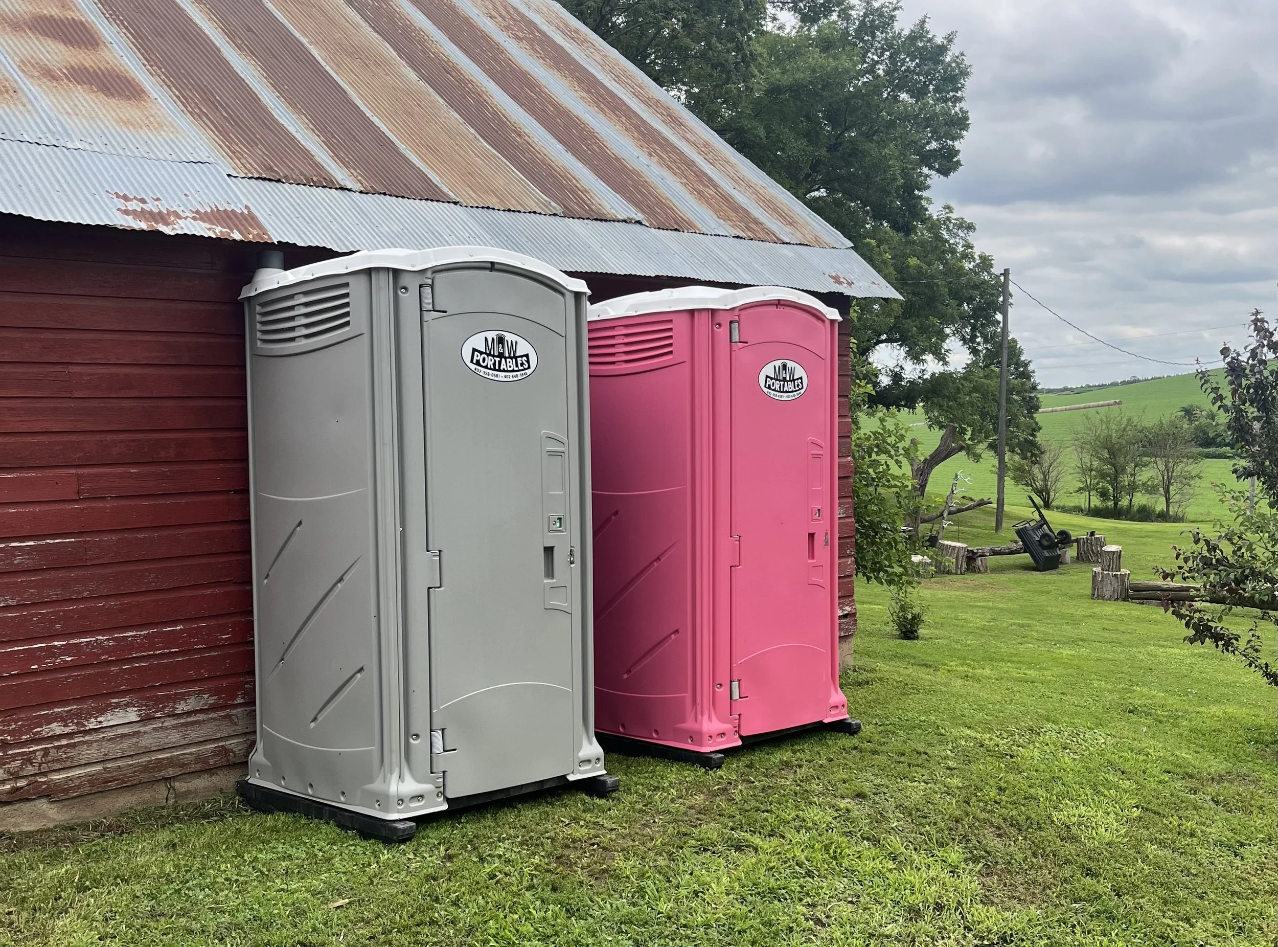 Two portable toilets, one gray and one pink, standing outside a red wooden building on a grassy area with trees and a green field in the background.