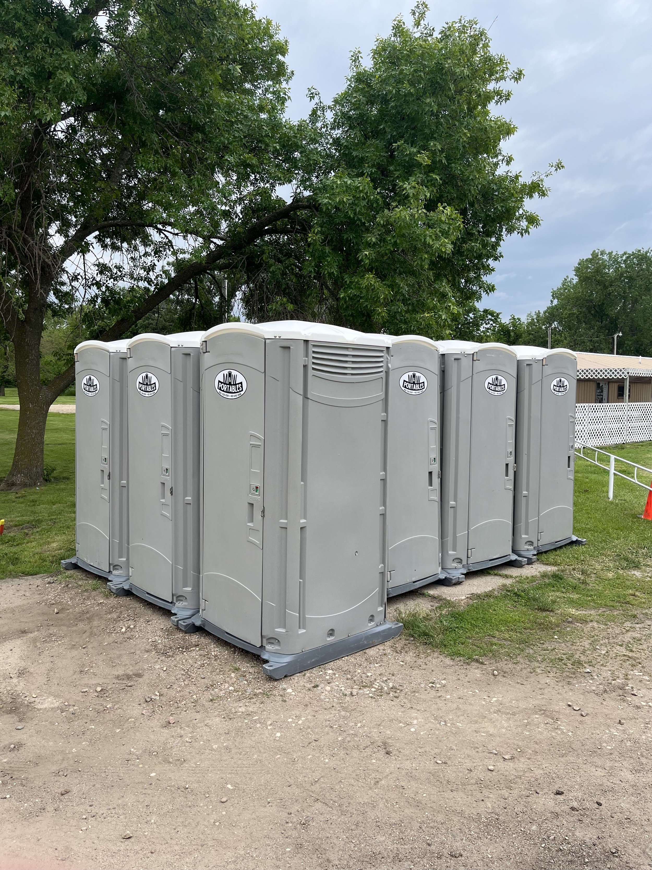 Six portable toilets arranged in a row outdoors on grass and dirt, with green trees and a building in the background.