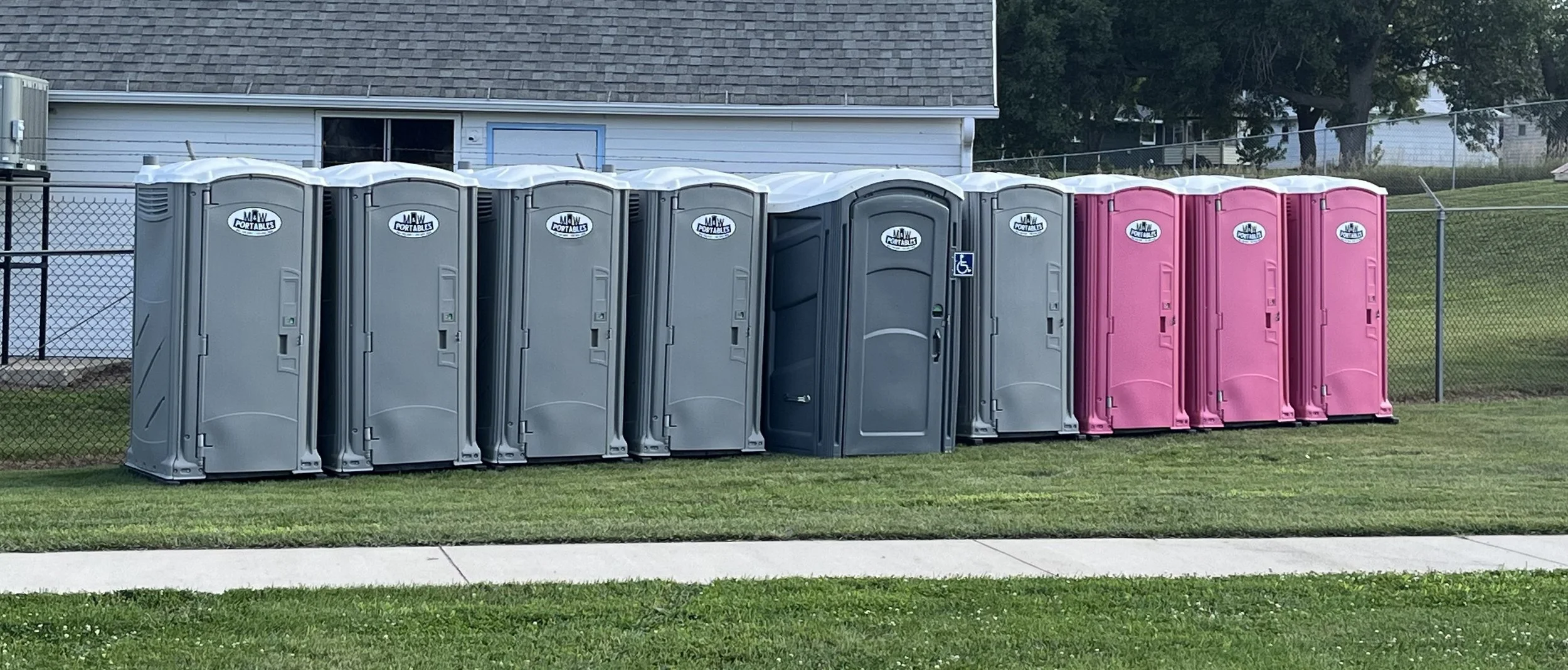Line of portable toilets, mostly gray with one pink, situated on a grassy area next to a sidewalk and a chain-link fence, with a house and trees in the background.