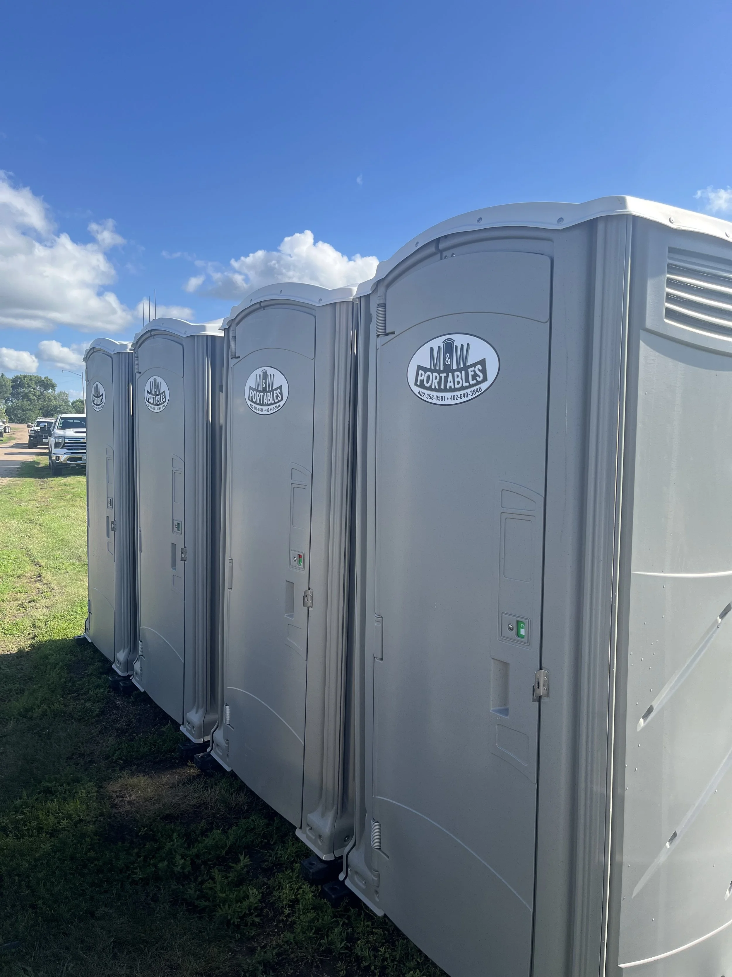 Four portable toilets lined up outdoors on grass with a clear blue sky and some white clouds in the background.