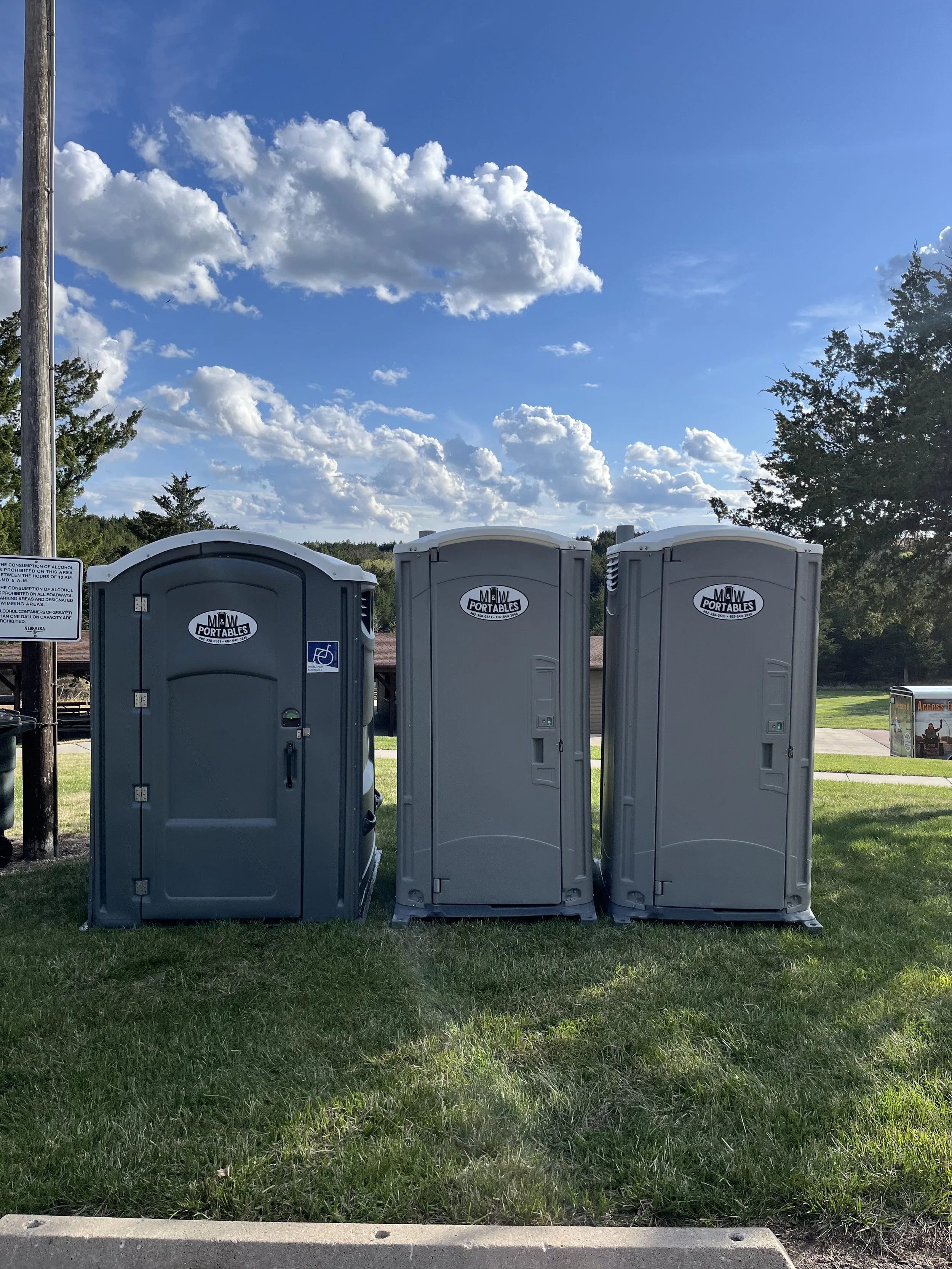 Three portable toilets are standing side by side on a grassy area with trees and a clear blue sky with scattered clouds in the background.
