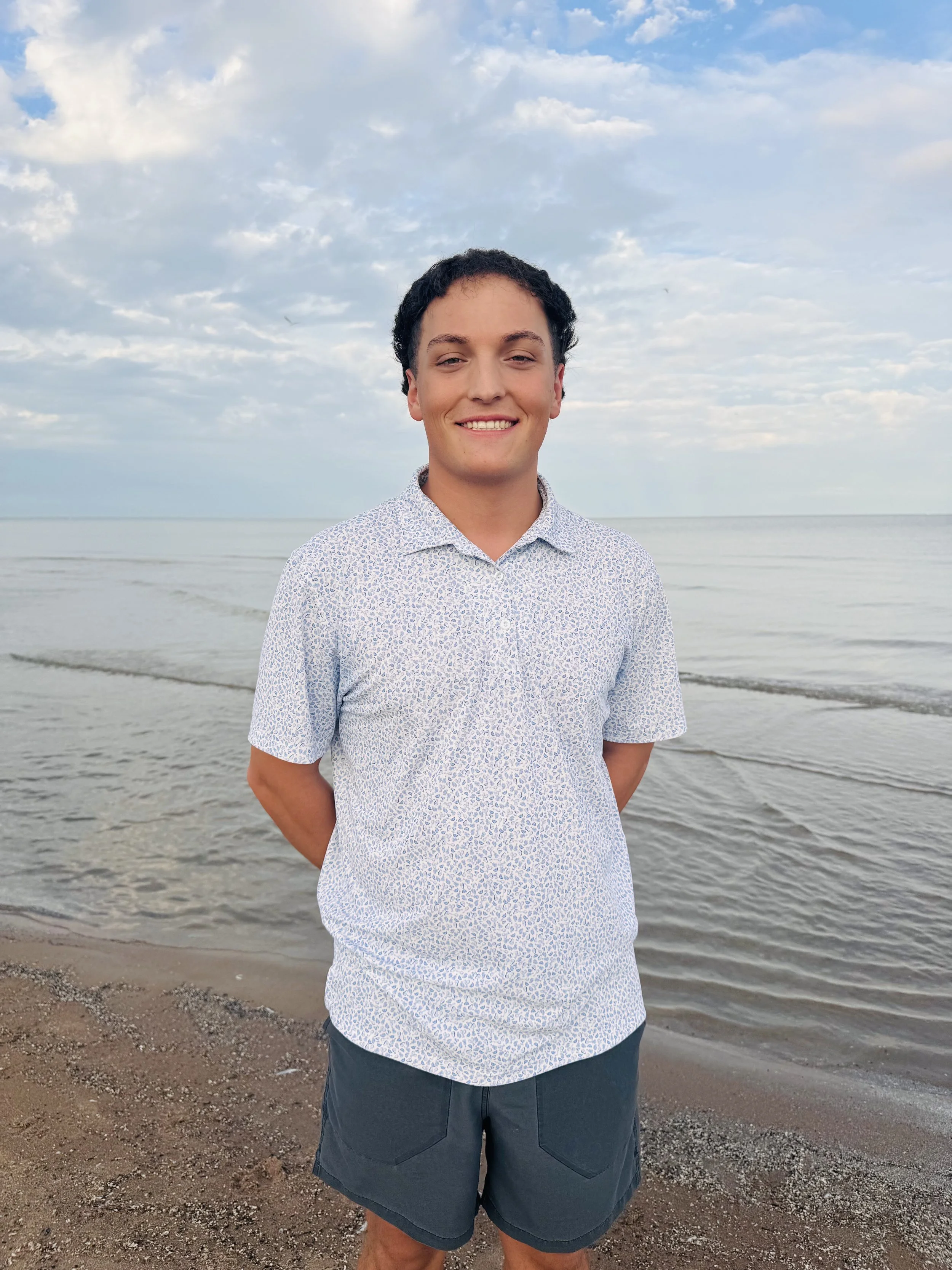 A young man standing on a sandy beach with water and a cloudy sky in the background. He's smiling, wearing a light patterned button-up shirt and dark shorts.