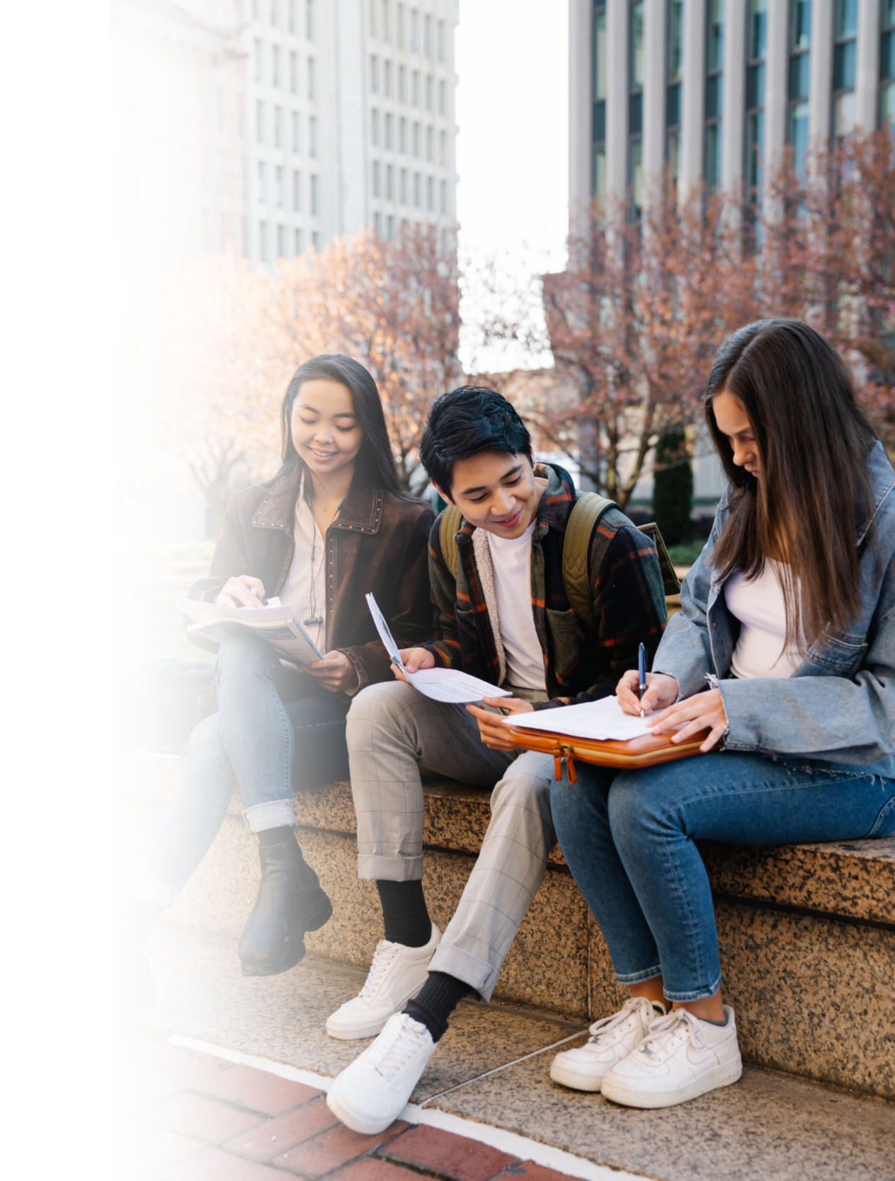 Three students sitting on a stone bench outdoors, studying and sharing notes with tall buildings and autumn trees in the background.