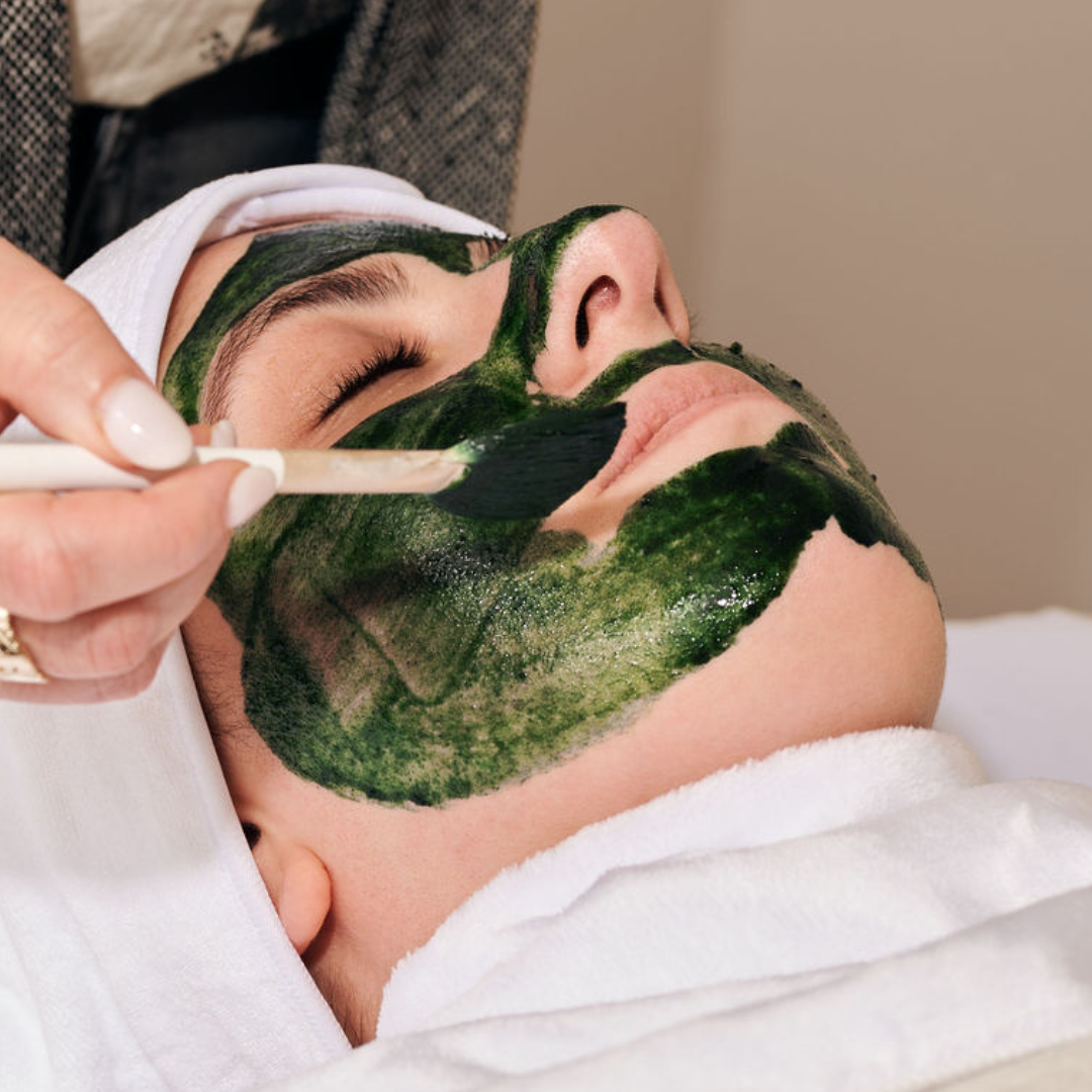 A woman receives a facial treatment with a green algae mask applied to her face, lying with eyes closed during the procedure.