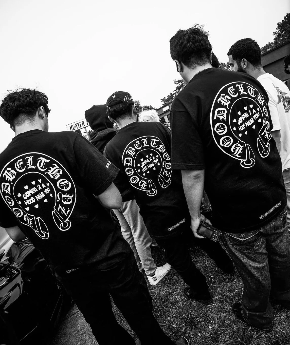 Group of young men wearing matching black t-shirts with a circular logo on the back, standing outdoors, viewed from behind in black and white.