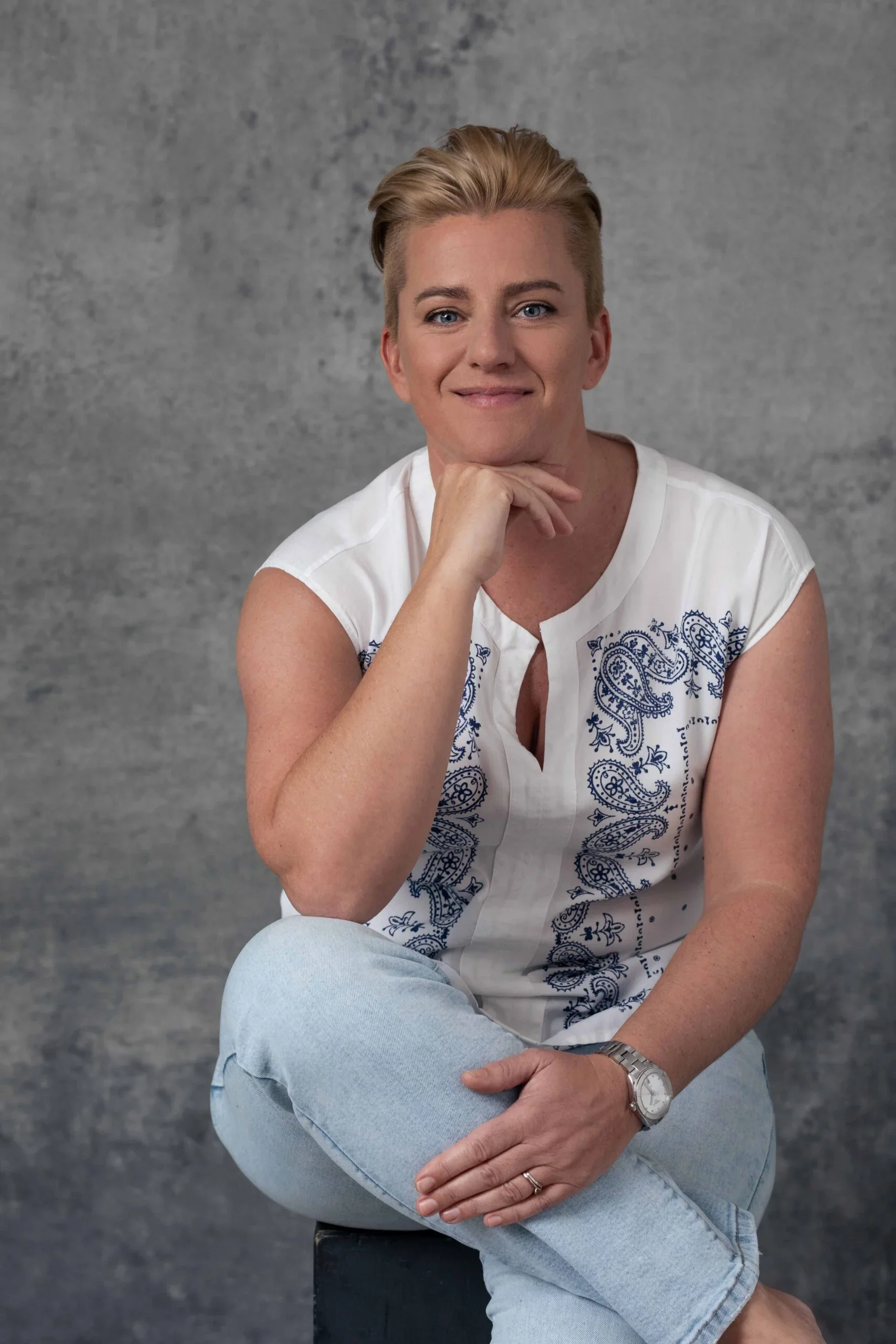 A woman with short blonde hair, wearing a white sleeveless top with blue embroidery, light blue jeans, and a watch, sitting on a black stool with her left leg crossed over her right, resting her chin on her right hand, in front of a gray textured background.