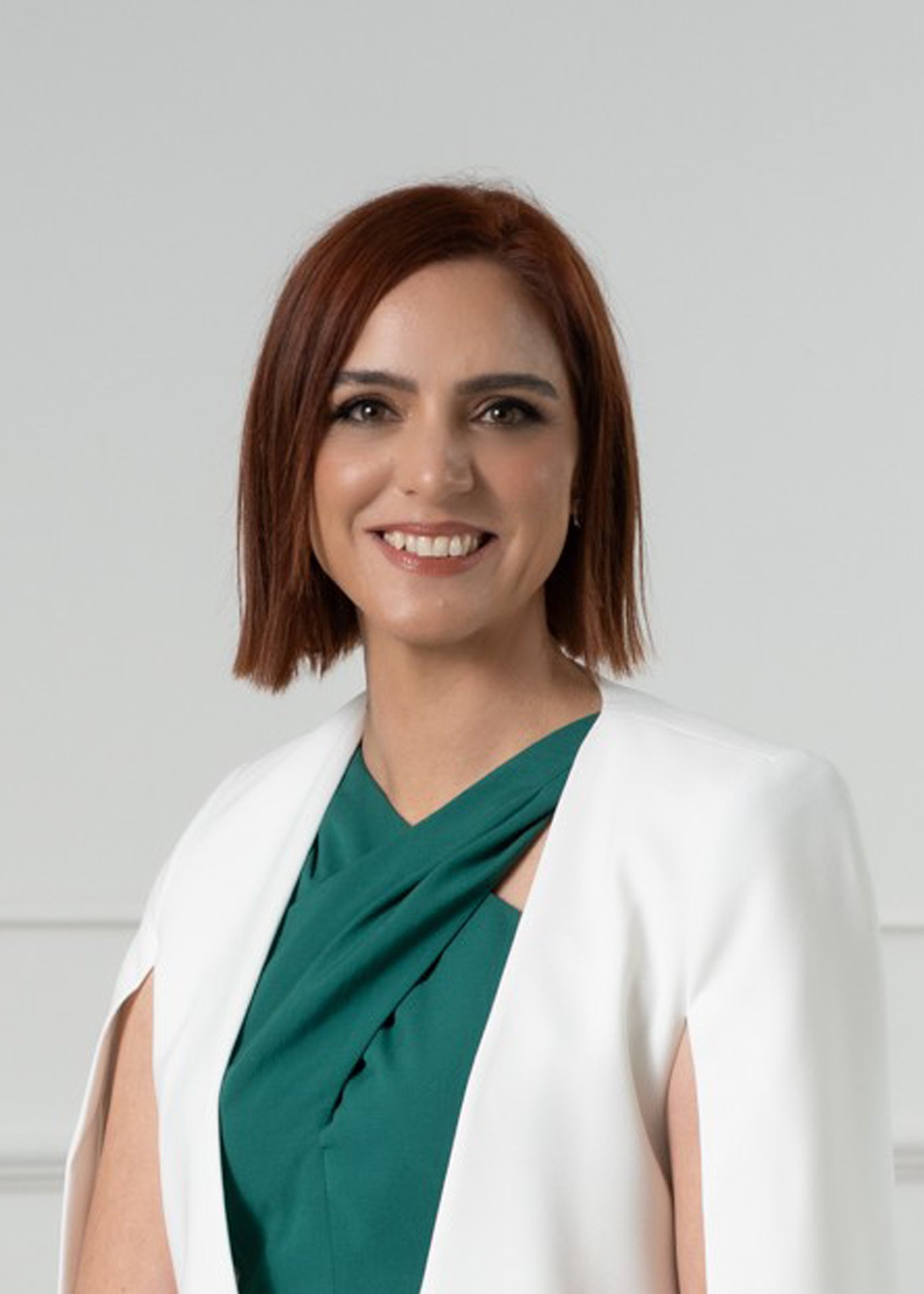 Portrait of a woman with short red hair, wearing a green top and white blazer, smiling, against a plain light background.