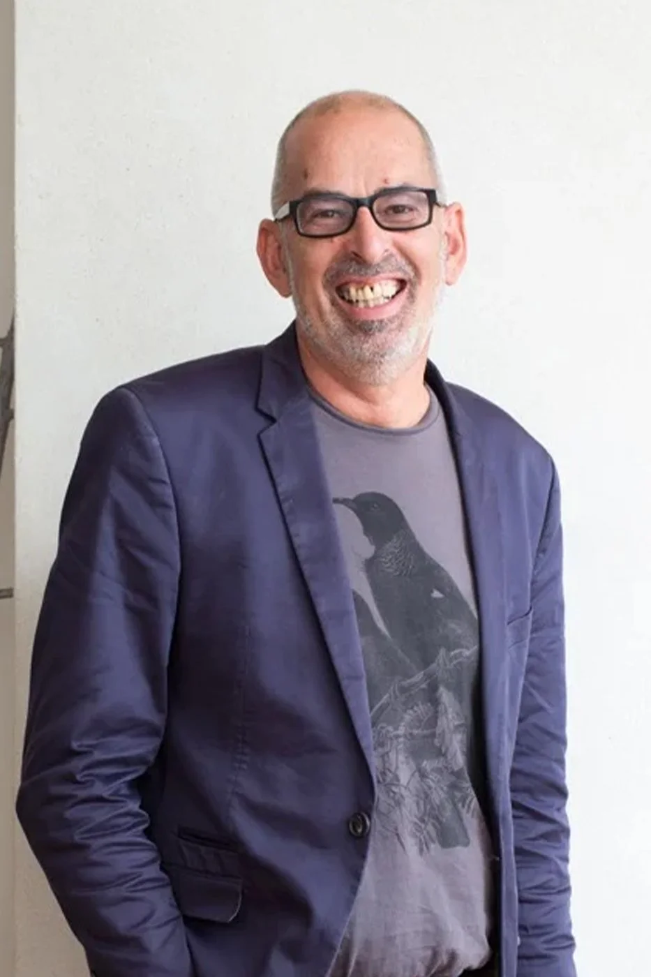 Man with glasses smiling, wearing a graphic T-shirt and a dark blazer, standing against a plain wall.