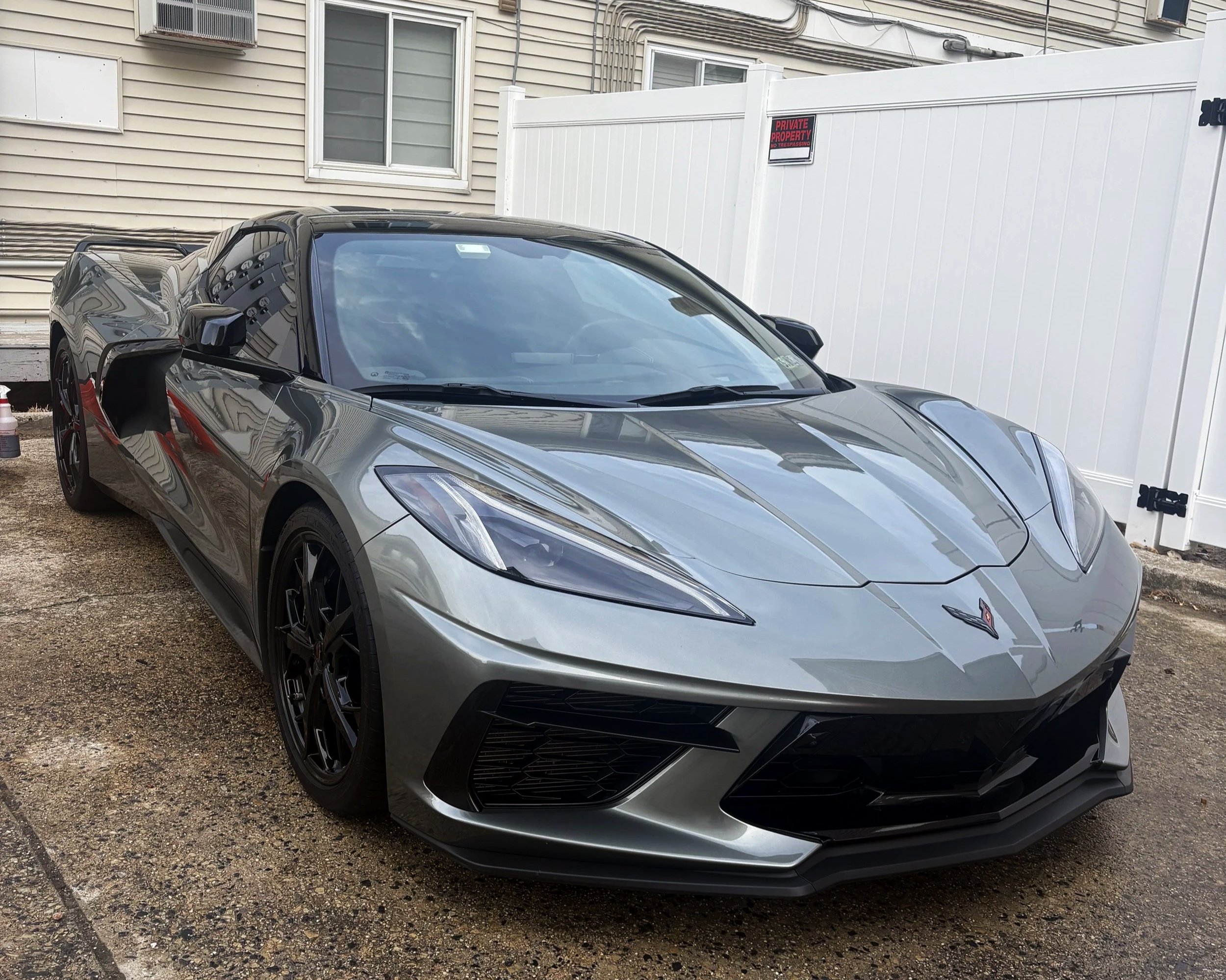 A silver Chevrolet Corvette sports car parked on a gravel surface near a white fence and beige house, with a 'Private Property' sign in the background.
