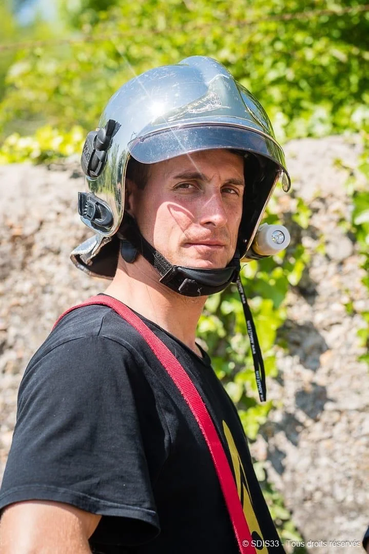 A man wearing a shiny motorcycle helmet with a sun glare reflection, black t-shirt, and red strap, standing outdoors with green foliage and rocks in the background.