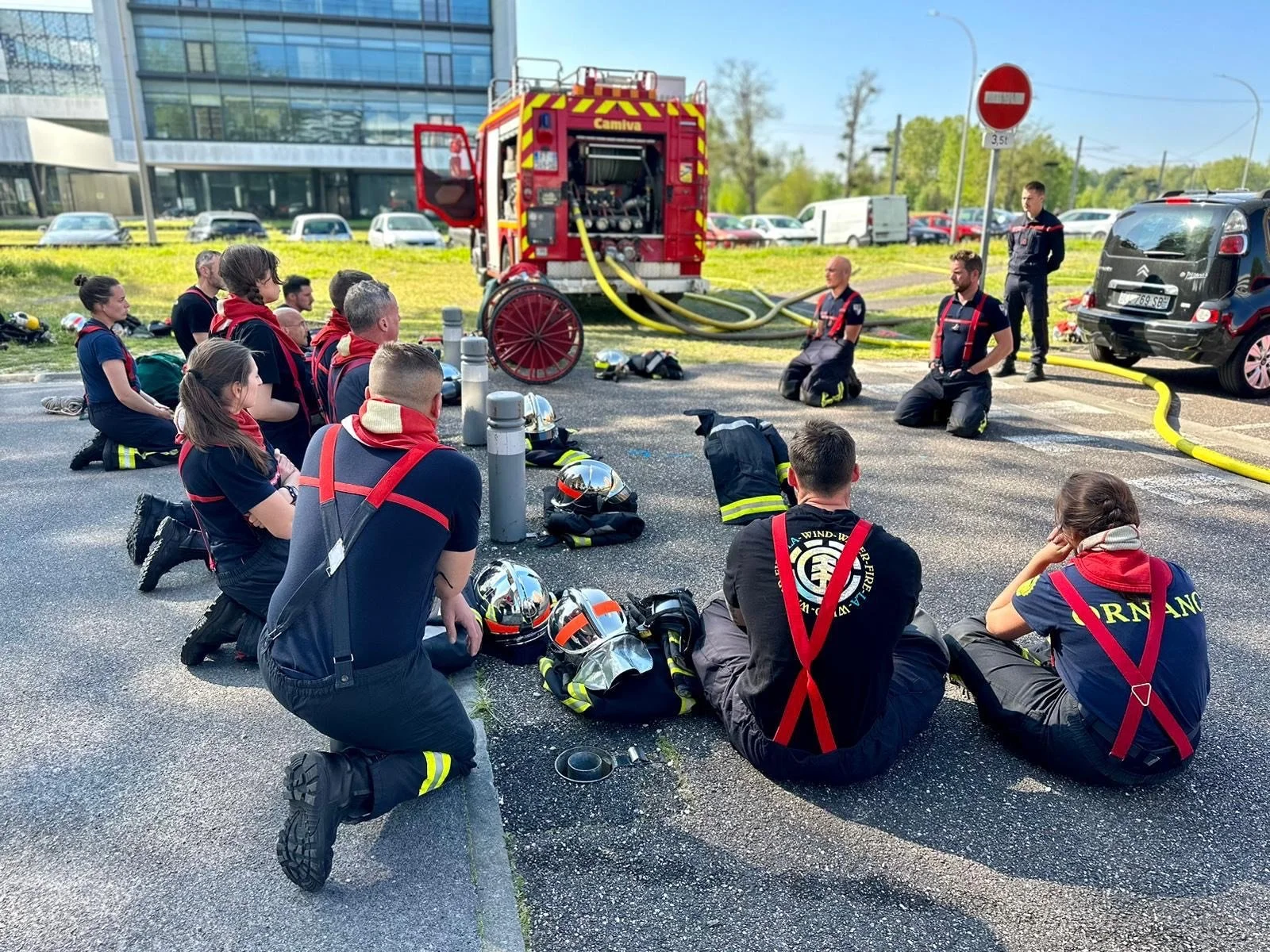 Firefighters in uniform kneeling on the ground during a safety training or briefing session outside. Fire hoses are connected to a large red fire pump. Helmets and other firefighting gear are placed on the ground nearby. Several cars are parked along the road, and a modern glass building is in the background. A no entry sign is visible, and the weather appears sunny and clear.