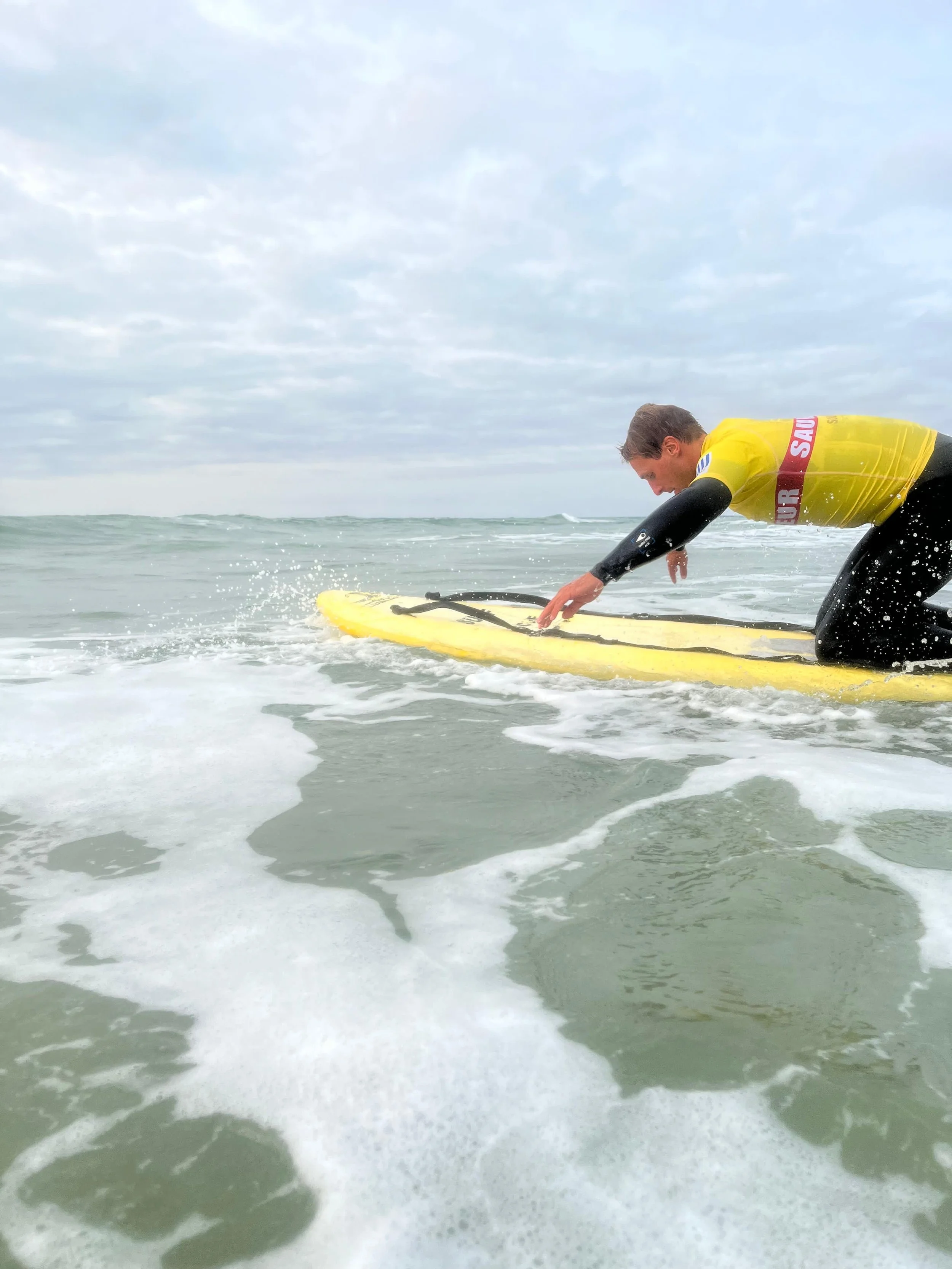 A man in a yellow shirt kneeling on a yellow surfboard in the ocean, preparing to stand up as waves surround him under a cloudy sky.