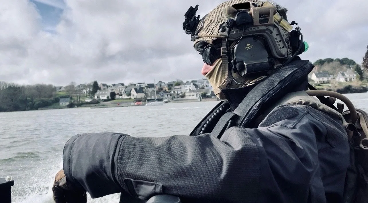 A person in military gear, including a helmet with mounted electronics, sunglasses, a face covering, and a tactical jacket, journeying on a boat with a water body and a town in the background.