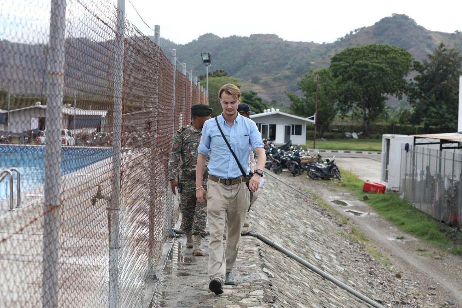 Three men walking on a narrow stone path beside a swimming pool, fenced with chain-link fence, with hills and trees in the background.