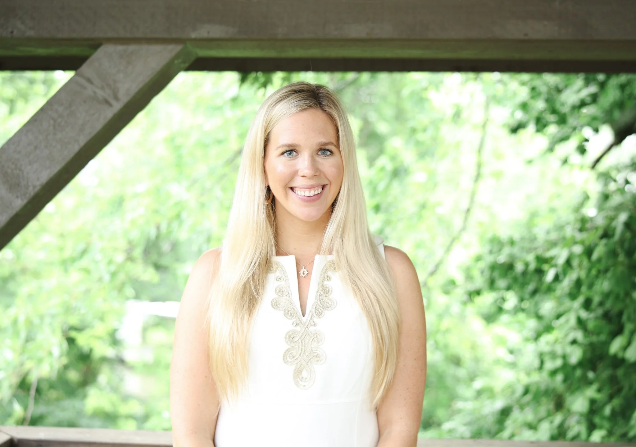 A young woman with long blonde hair smiling outdoors under a wooden structure with lush green foliage in the background.