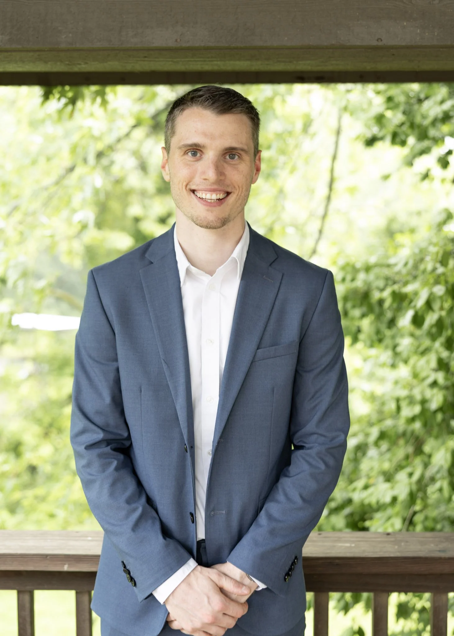 Young man in a blue suit and white shirt smiling outdoors on a porch with green trees in the background.