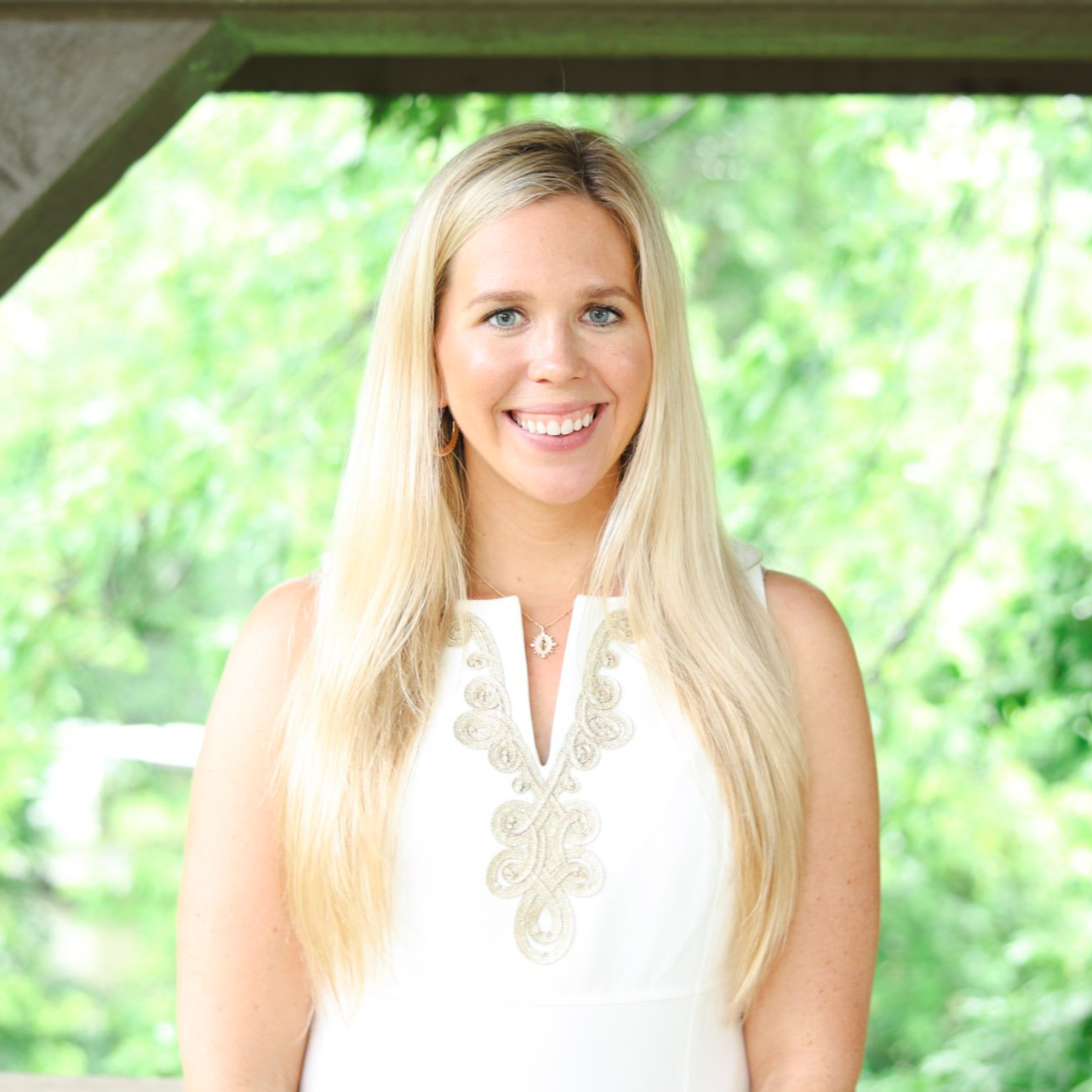 A smiling woman with long blonde hair wearing a white sleeveless dress with decorative embroidery, standing outdoors with a green leafy background.
