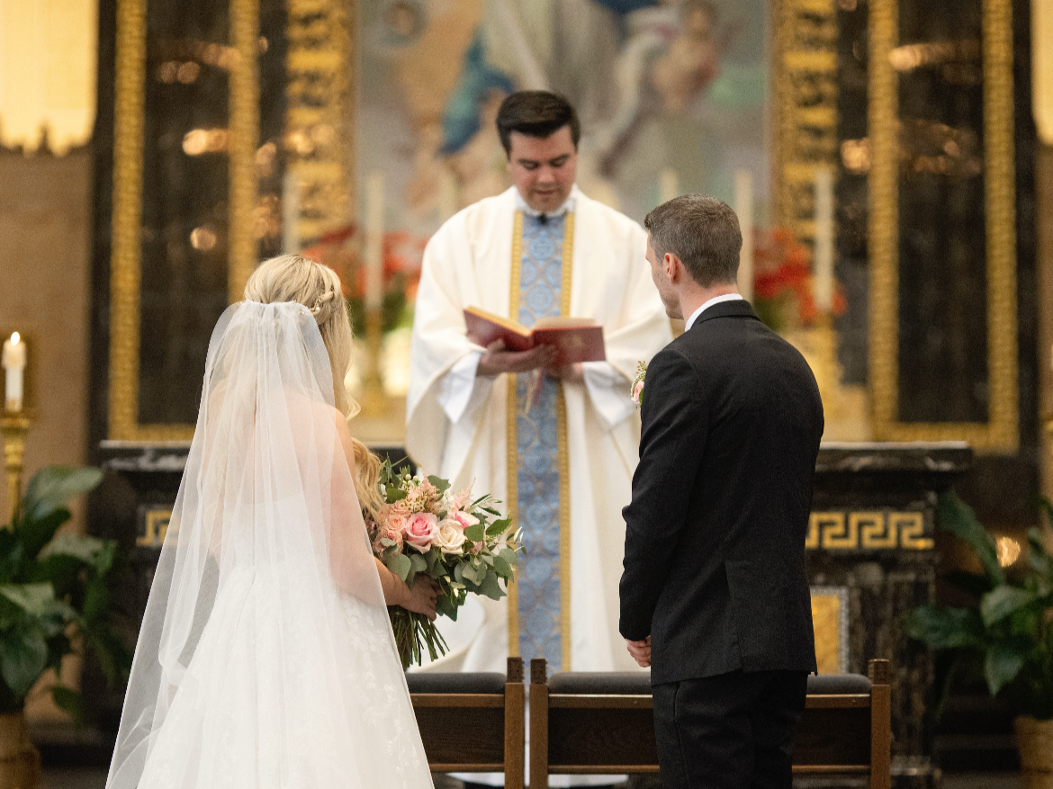 A bride and groom standing before a priest during a wedding ceremony inside a church, with the bride holding a bouquet of pink roses and facing the priest.