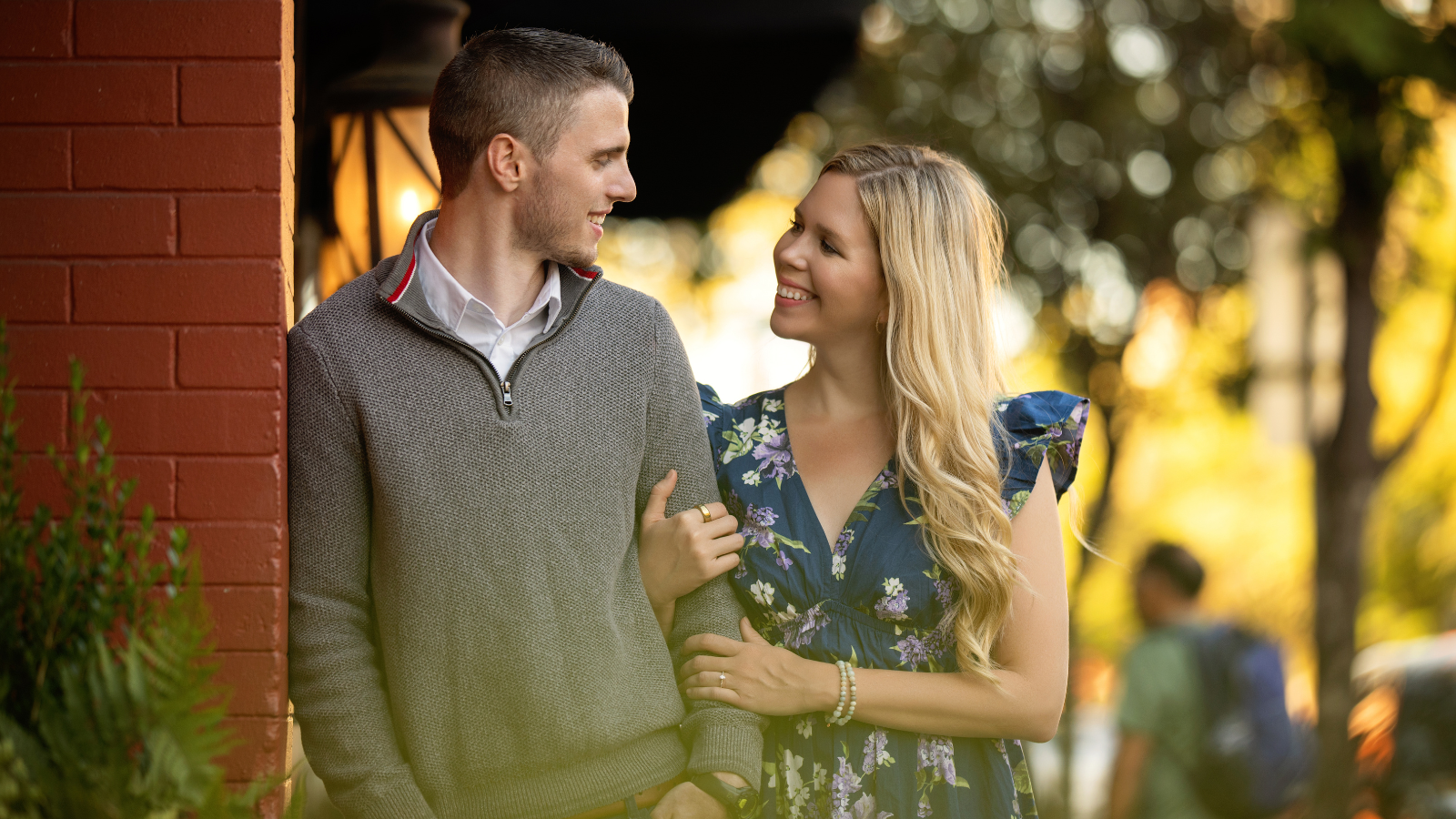 A young man and woman standing near a brick wall outdoors in warm sunlight, smiling at each other, the woman with her hand on the man's arm.