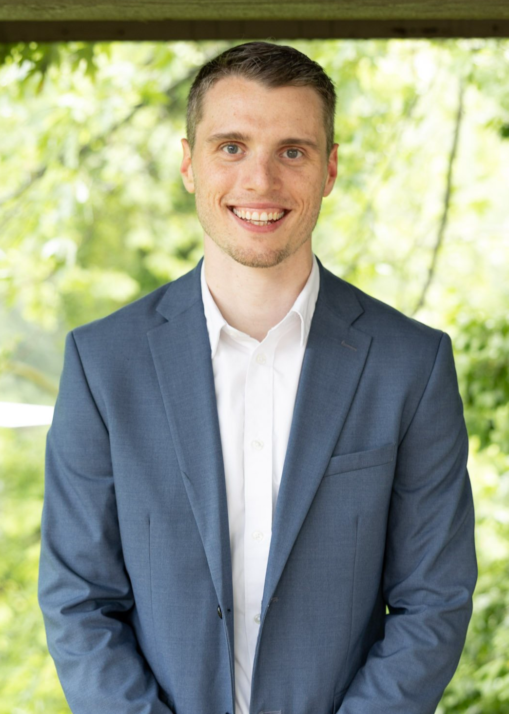 A young man with short brown hair, wearing a blue suit and white shirt, smiling at the camera outdoors with blurred green trees in the background.