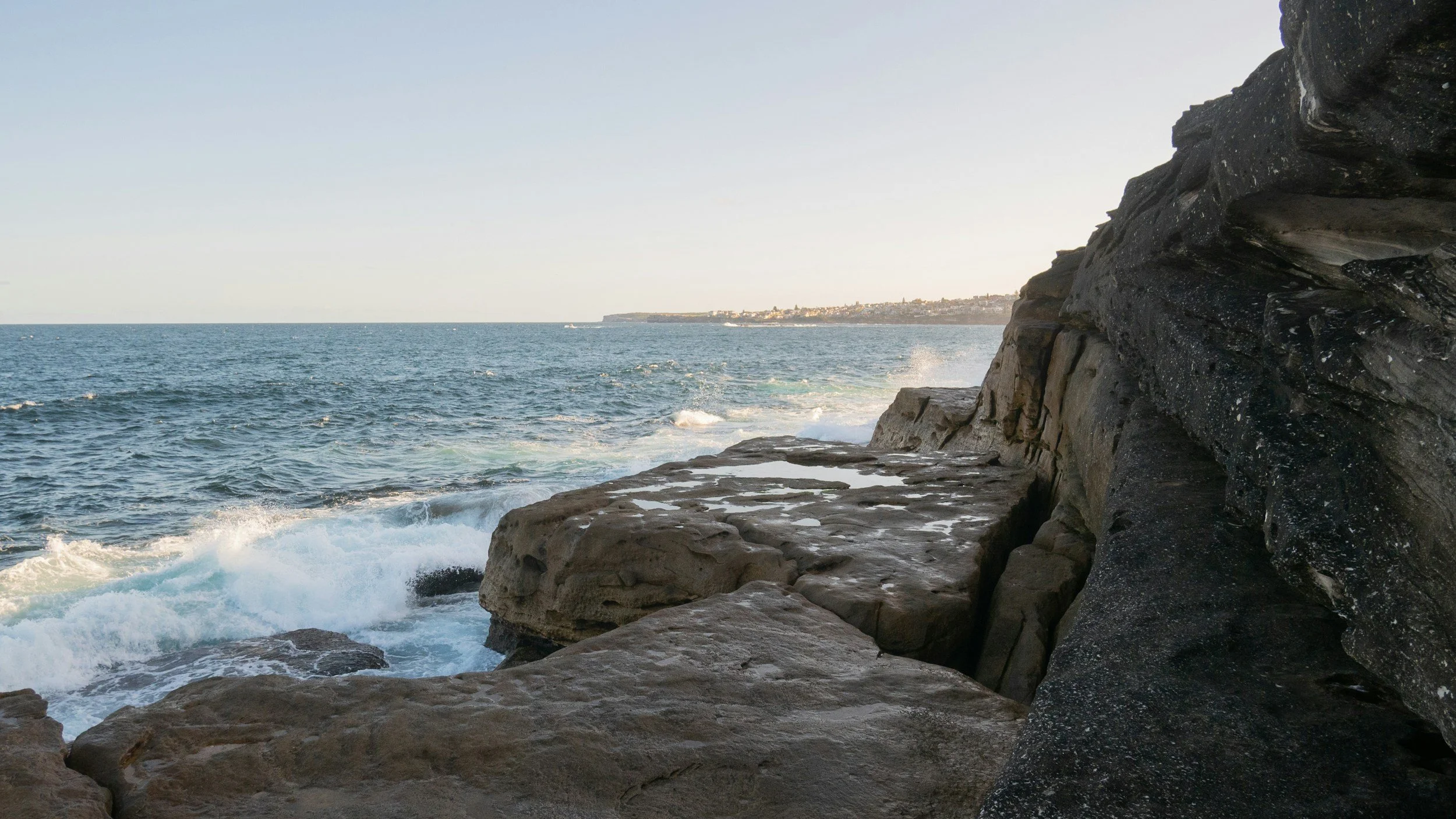 Rocky coastline with waves crashing against the shore, the ocean extends to the horizon with a distant landmass.