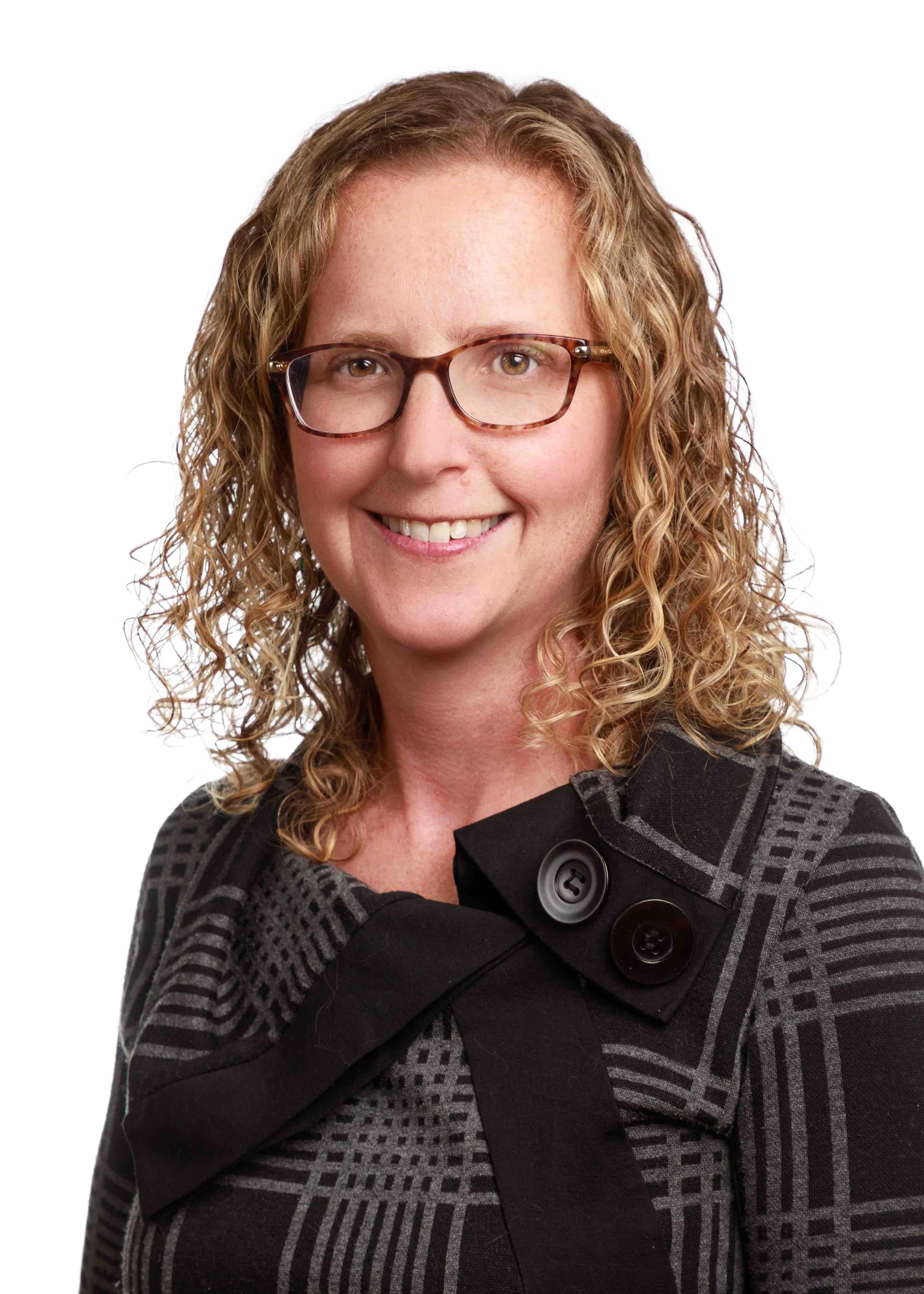 A woman with curly blonde hair and glasses smiling at the camera, wearing a patterned black and gray top with large black buttons.