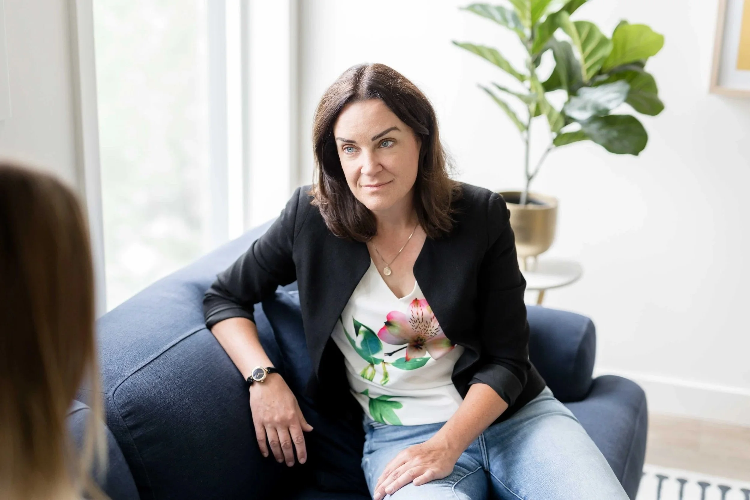 A woman with dark brown hair wearing a black blazer, white floral shirt, and light blue jeans, sitting on a blue couch during a conversation in a well-lit room with a large window and a potted plant in the background.