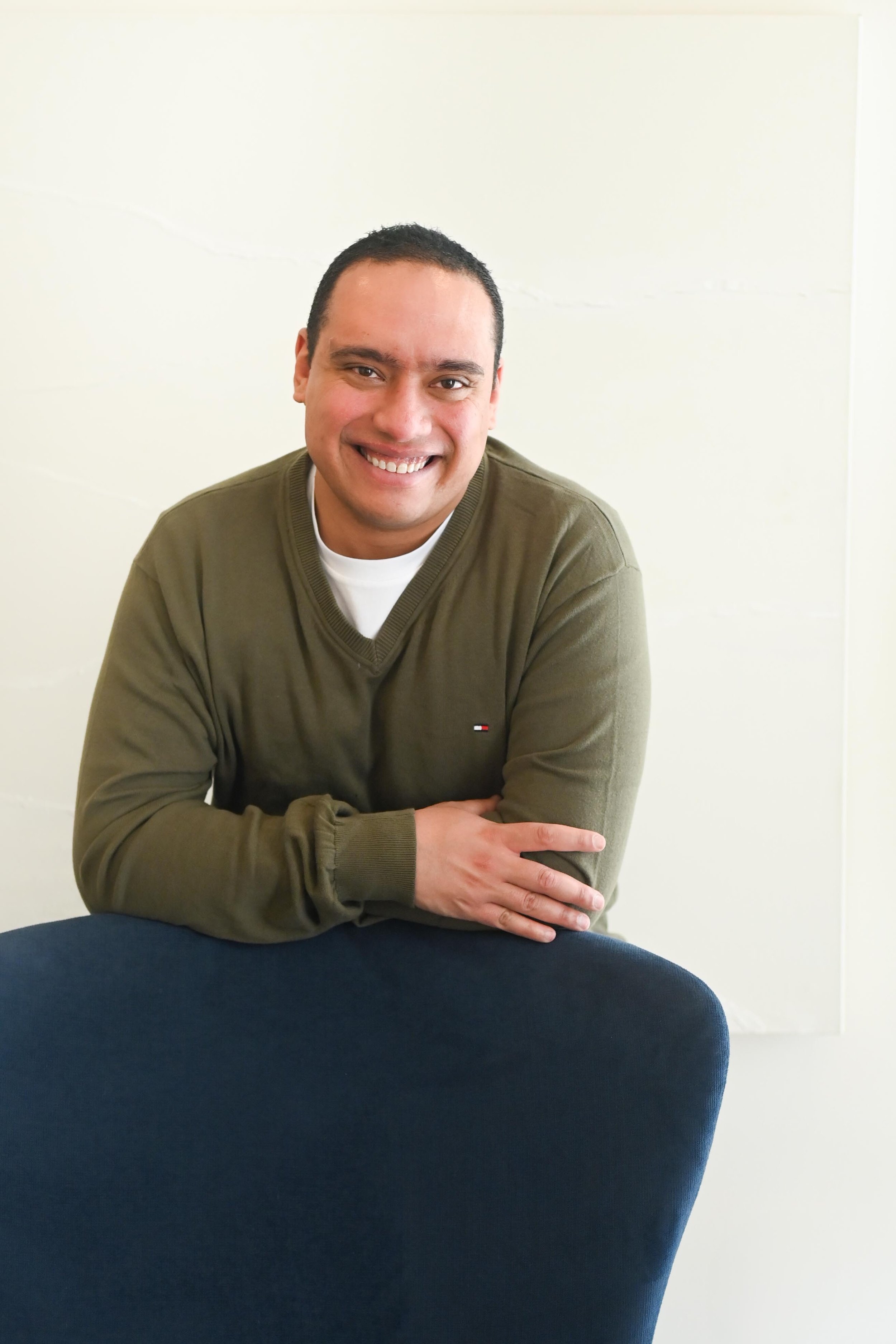 A smiling man with short dark hair, wearing an olive green sweater and white shirt, leaning on a blue chair in an indoor setting with a plain light-colored wall background.