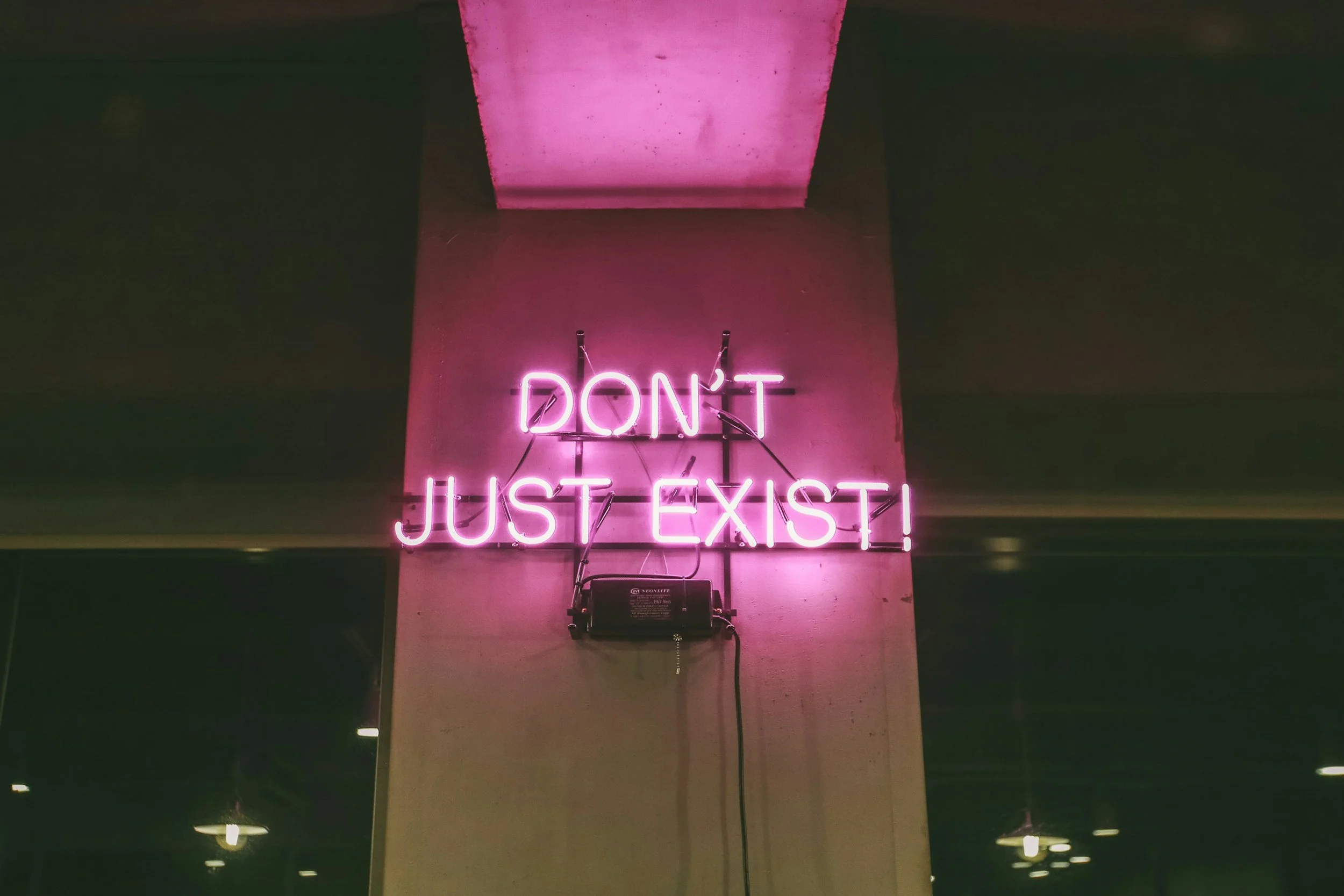 Pink neon sign on a wall that says "DON'T JUST EXIST!" in capital letters.