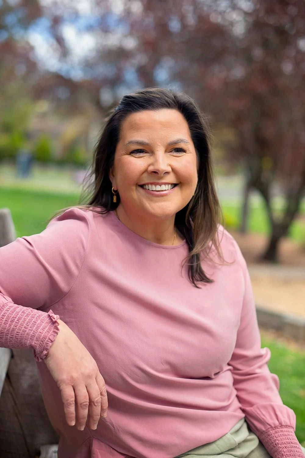 Crissi, smiling outdoors in a park, wearing a pink long-sleeve top with textured cuffs, with trees and greenery in the background.