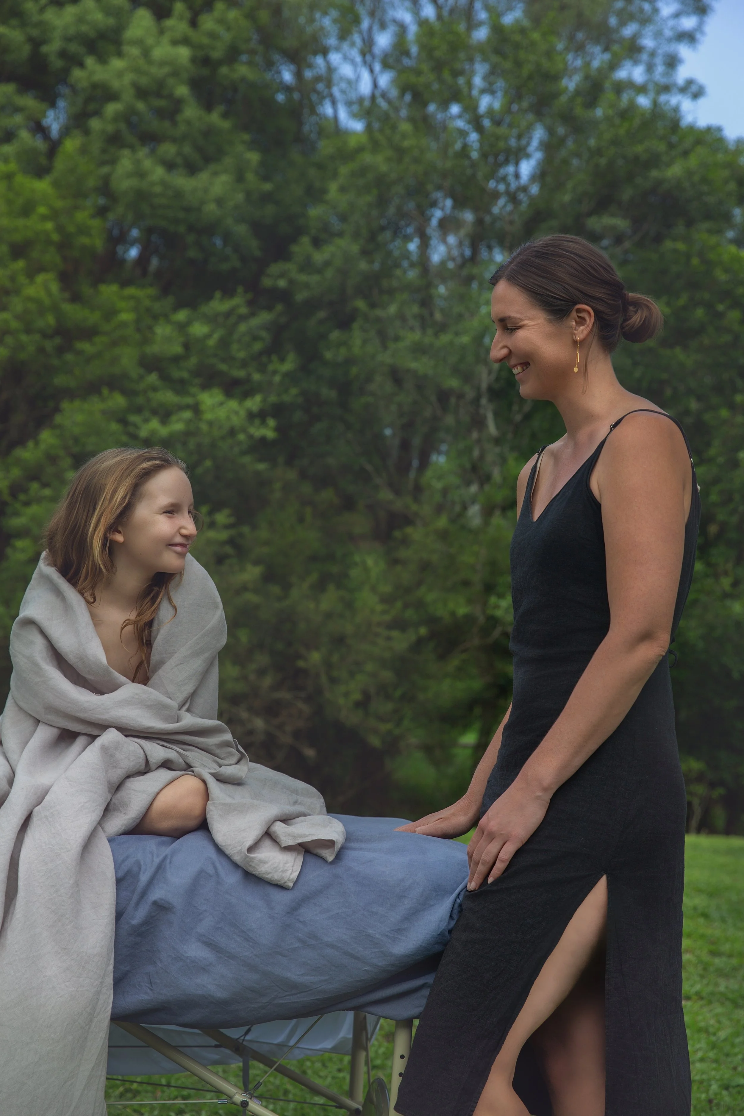 A woman in a black dress smiling at a girl wrapped in a beige blanket outdoors with green trees in the background.