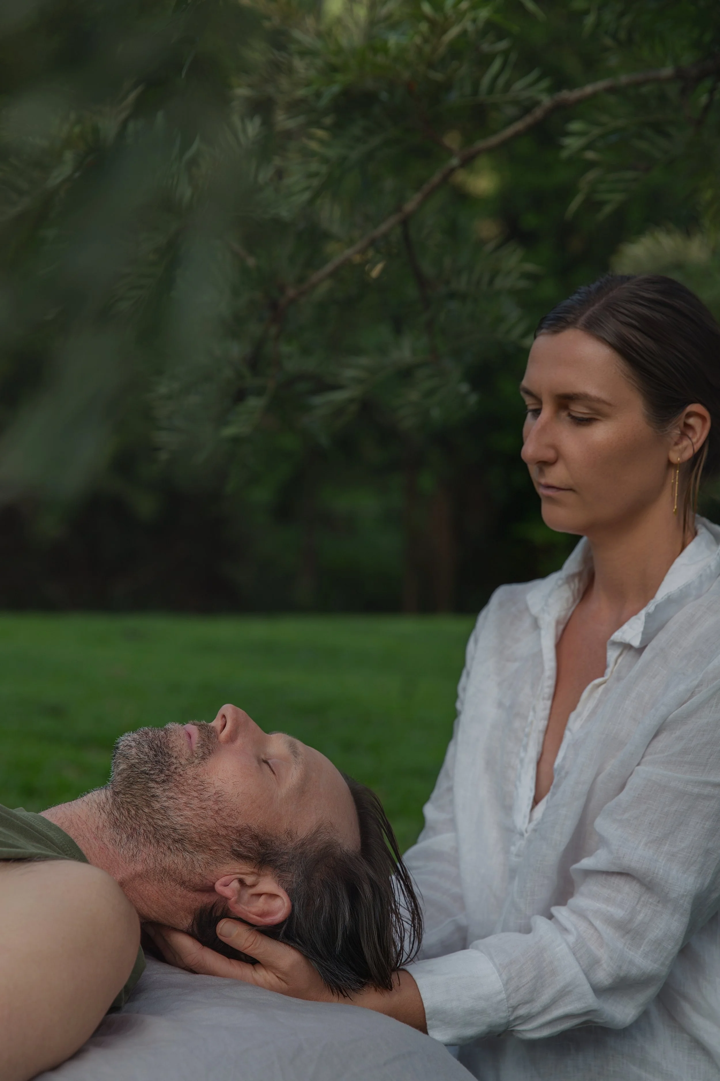 A woman is sitting outdoors in a park or garden with green grass and trees in the background. She is gently holding the head of a man and is offering craniosacral therapy for nervous system support. It is a moment of care and tenderness.