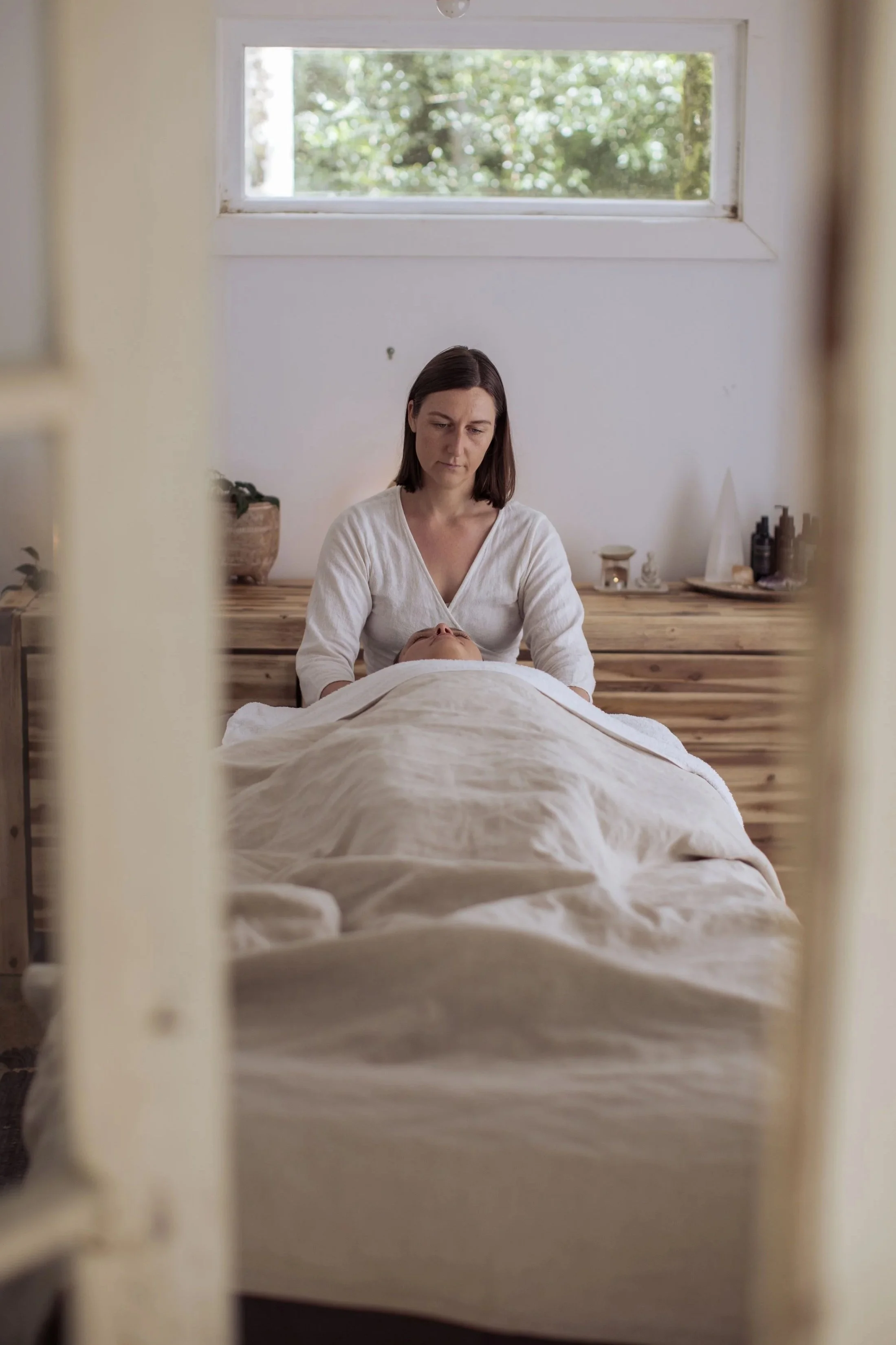 A woman lies on a bed covered with a beige blanket, viewed through a mirror frame, with another woman tending to her bedside in a room with a window and wooden furniture.