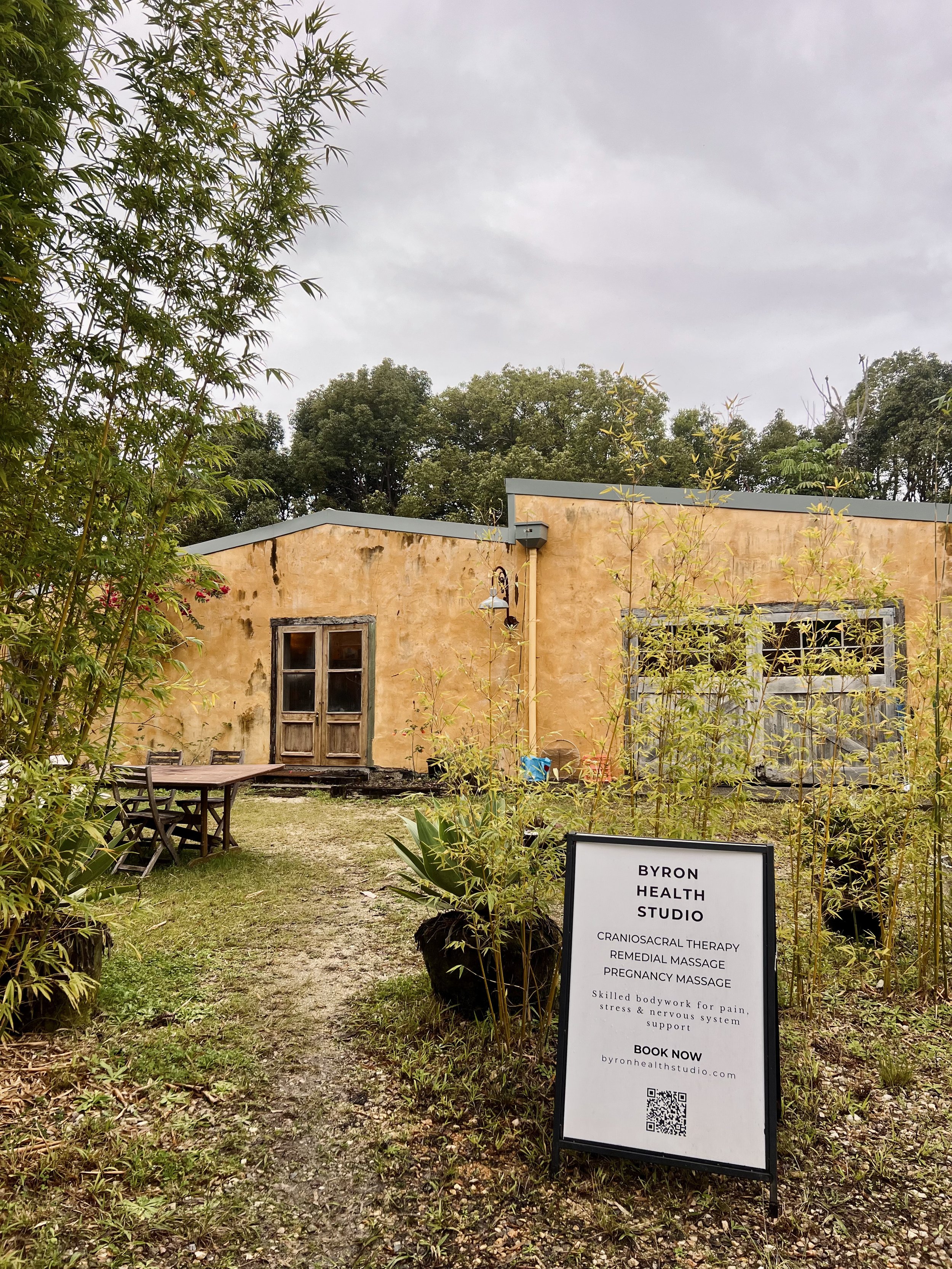 An outdoor scene with a rustic building, trees, and a signboard for Byron Health Studio offering craniosacral therapy, remedial massage, pregnancy massage, and other services, with overcast sky and some outdoor furniture.
