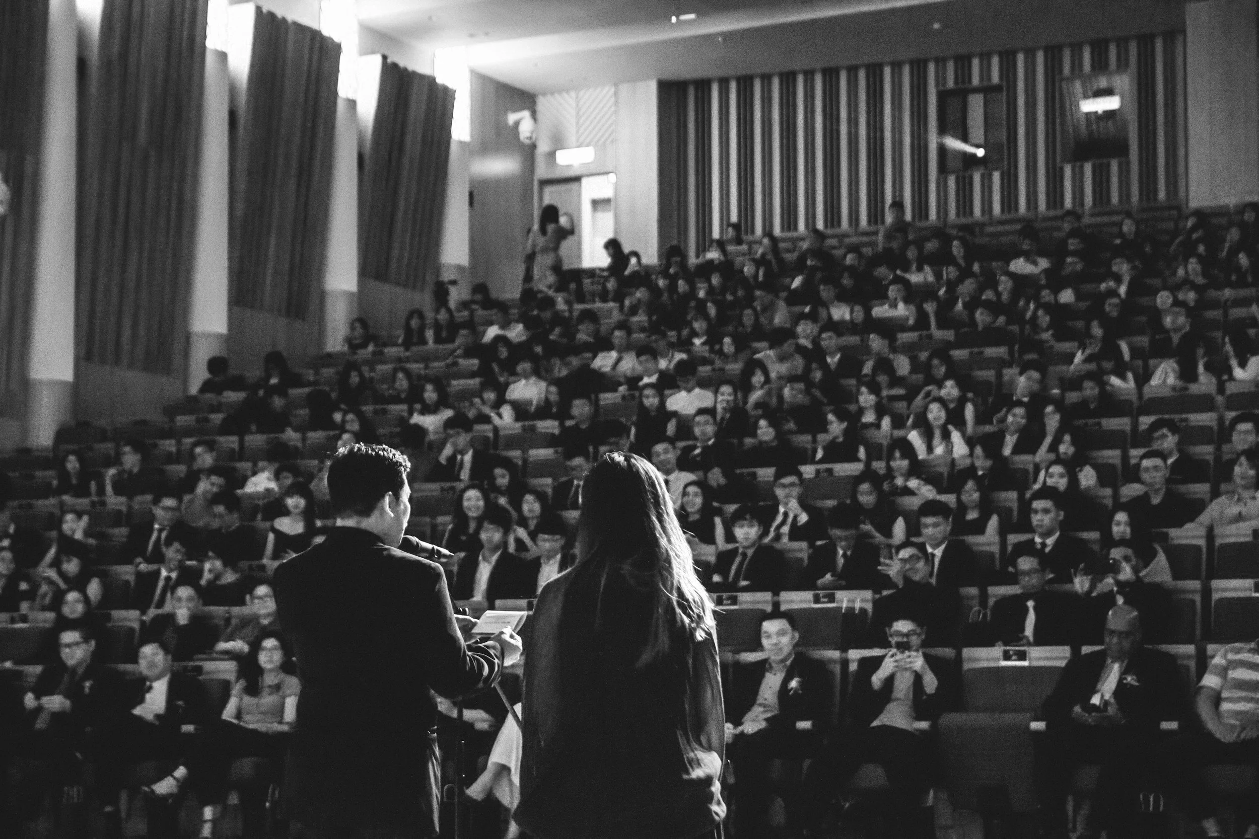 Una conferencia o presentación en un auditorio lleno de personas, con un hombre y una mujer en el escenario hablando frente a la audiencia.