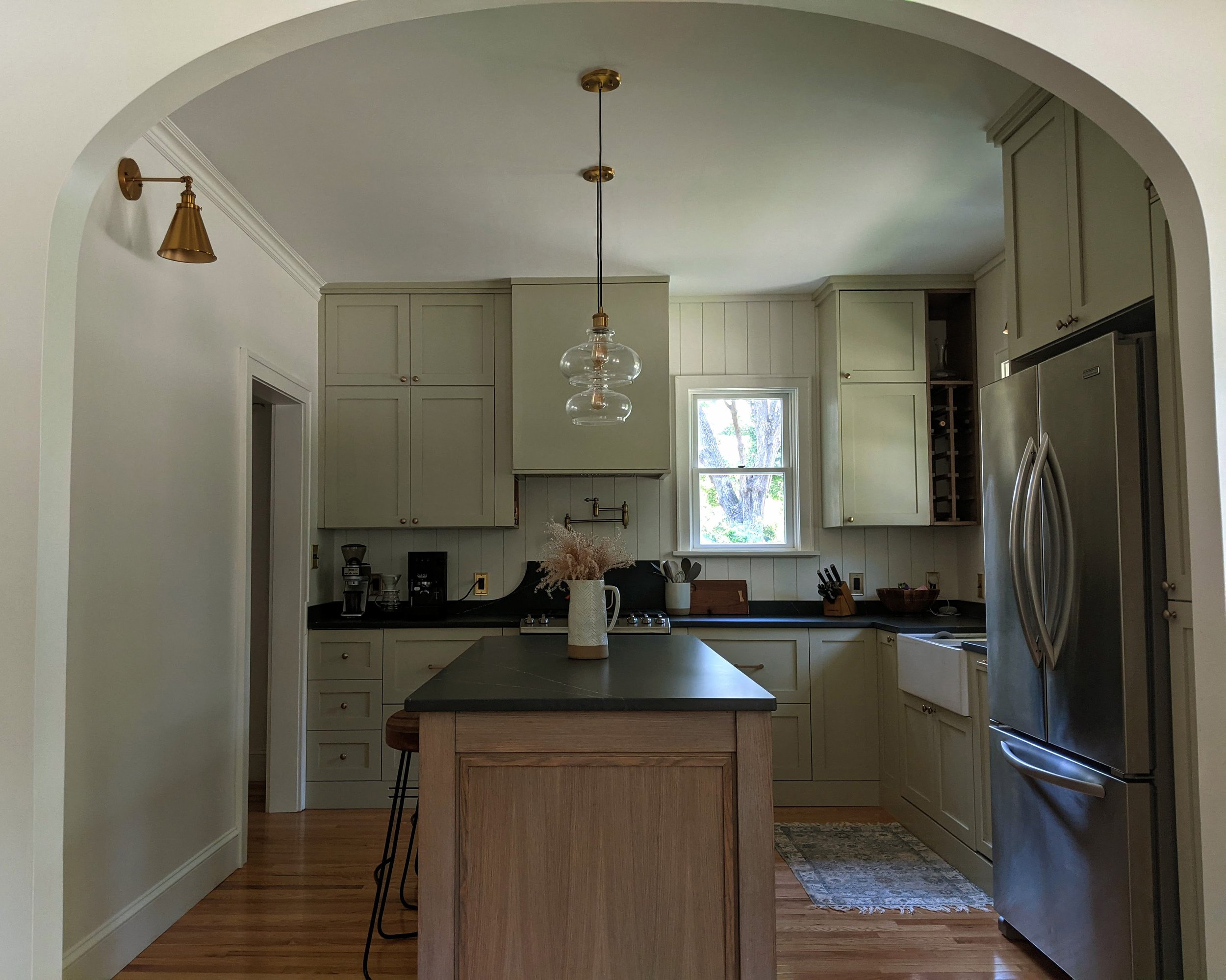 Kitchen with green cabinets, stainless steel refrigerator, wooden island, black countertops, window showing greenery outside, pendant light, and wall-mounted light fixture.