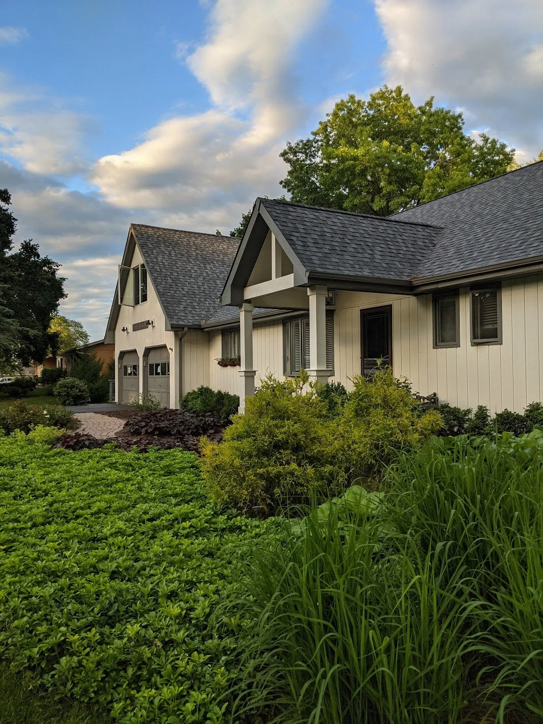 A modern house with a gabled roof, white exterior walls, and a front porch, surrounded by lush green landscaping and trees, under a partly cloudy sky.