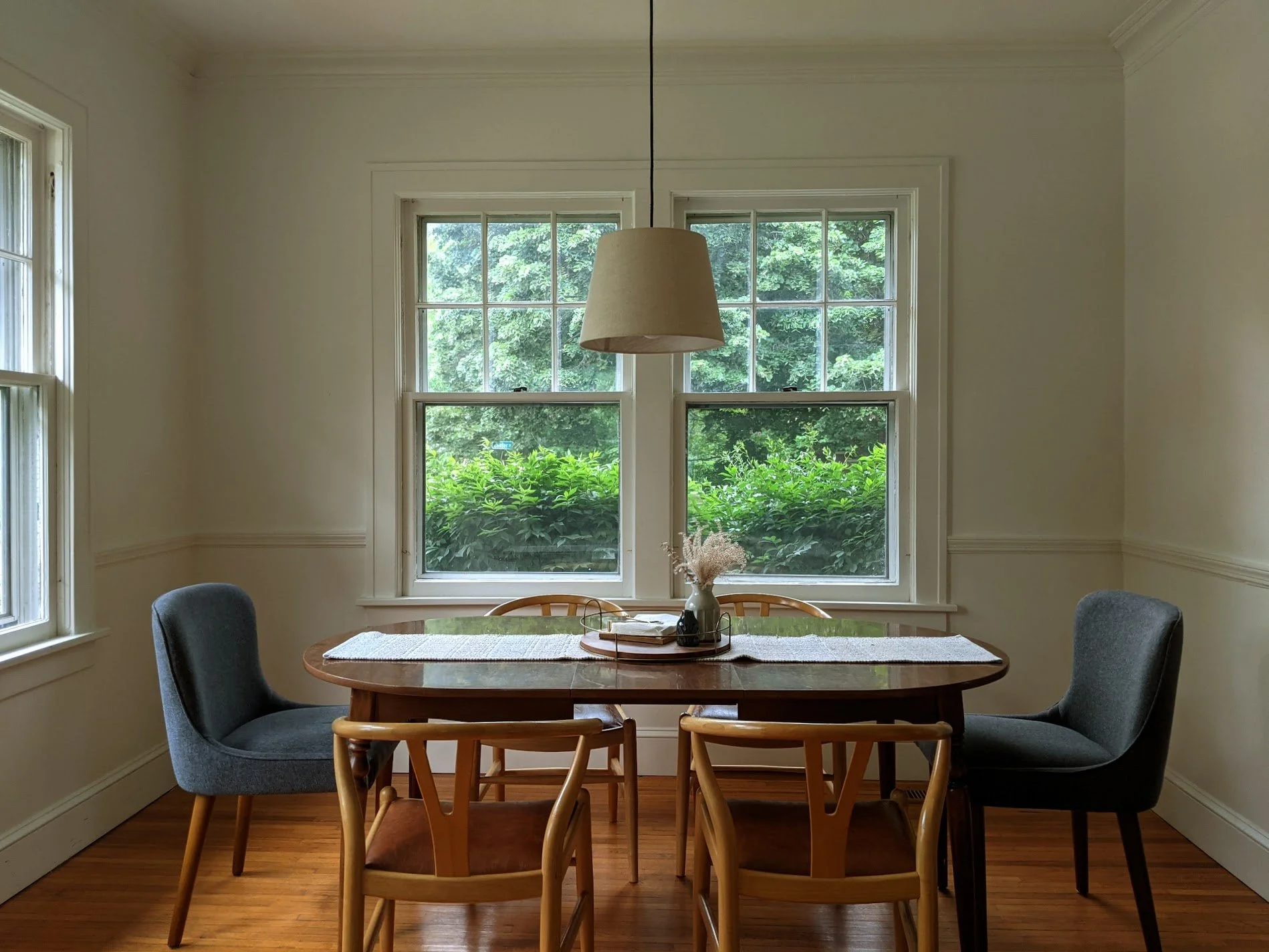 Dining room with wooden table, four chairs, and three windows, green foliage outside, white walls, and a ceiling lamp.