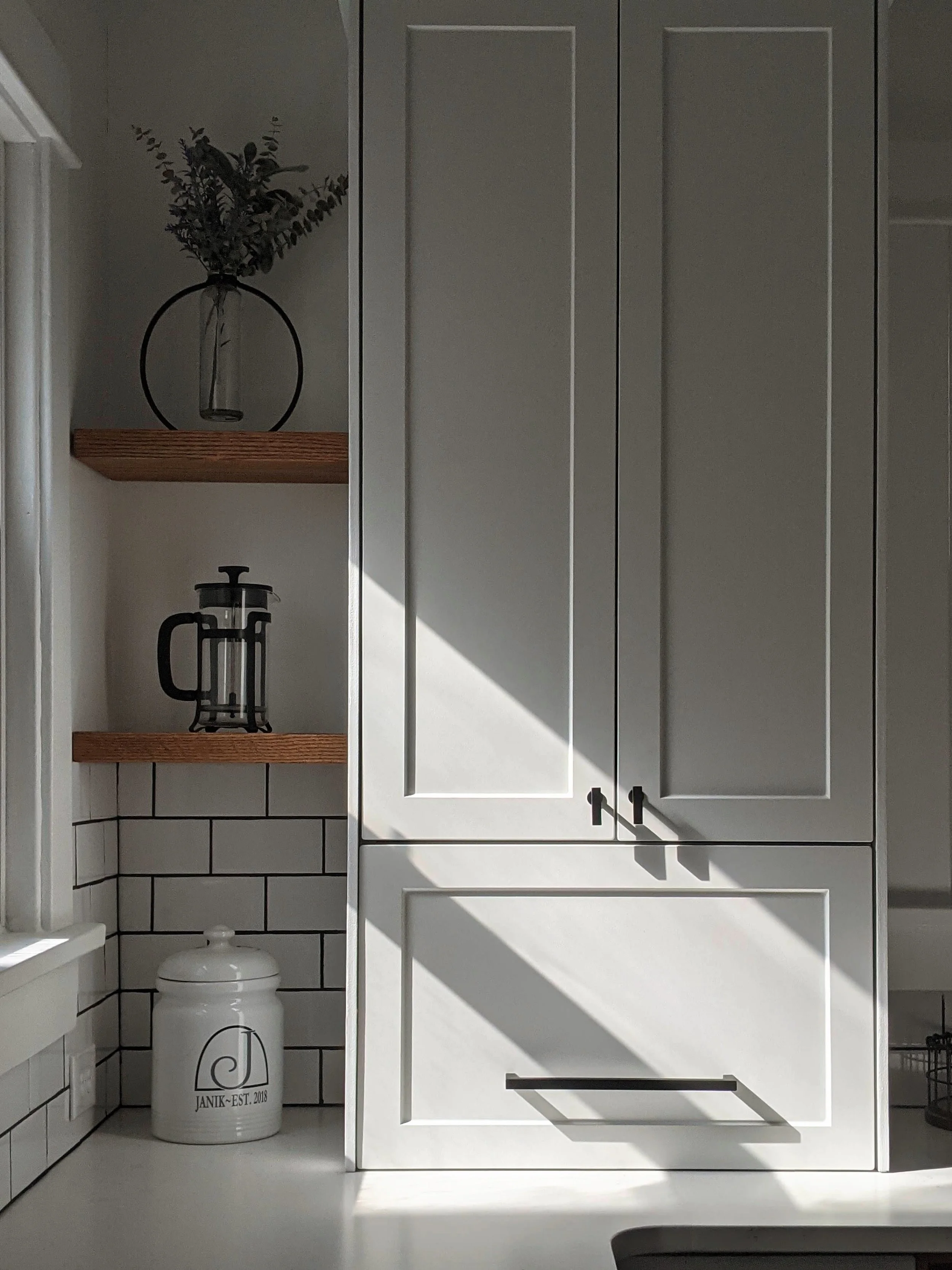 Kitchen cabinet with closed doors, shadowed area, white subway tile backsplash, open shelves with a glass mason jar with greenery and a French press coffee maker, white jar with black logo and text, and a black handle pull.