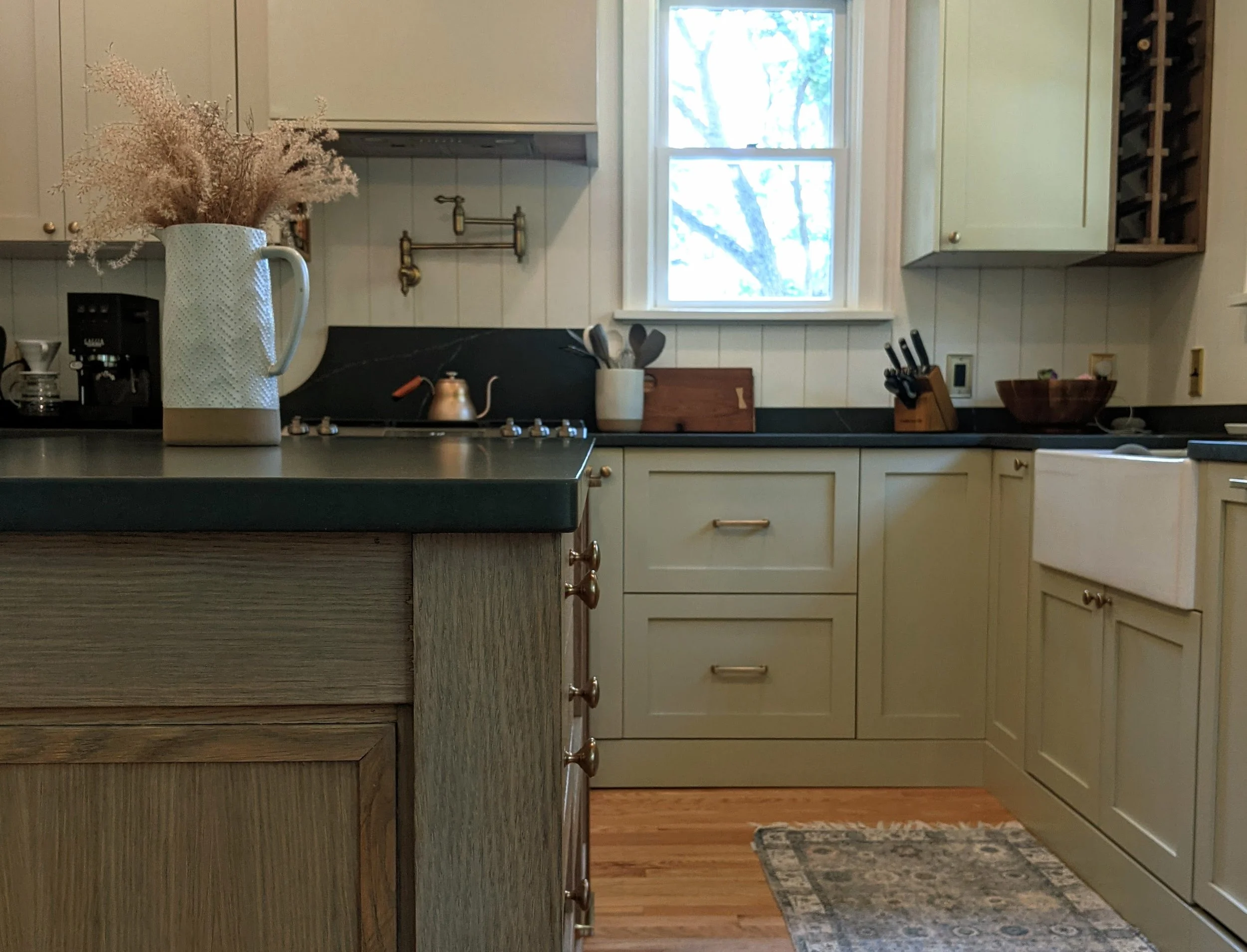 A cozy kitchen with cream-colored cabinets, black countertops, and a window showing trees outside. There is a large white pitcher with dried flowers, a coffee maker, a teapot on the stove, and various kitchen utensils on the counters.