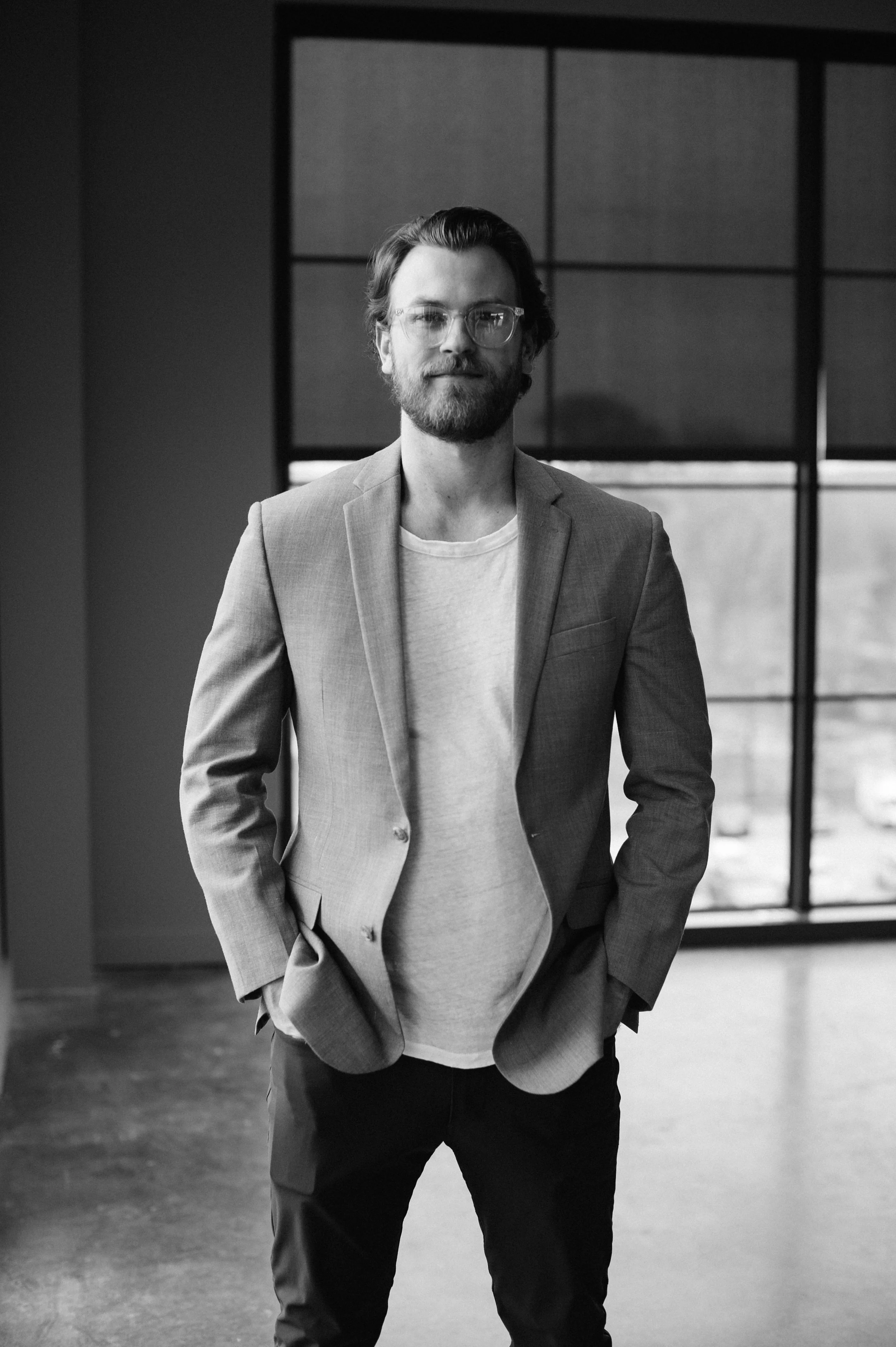 Black and white photograph of a man with glasses, a beard, and medium-length hair, wearing a casual blazer over a T-shirt, standing indoors with a large window in the background.