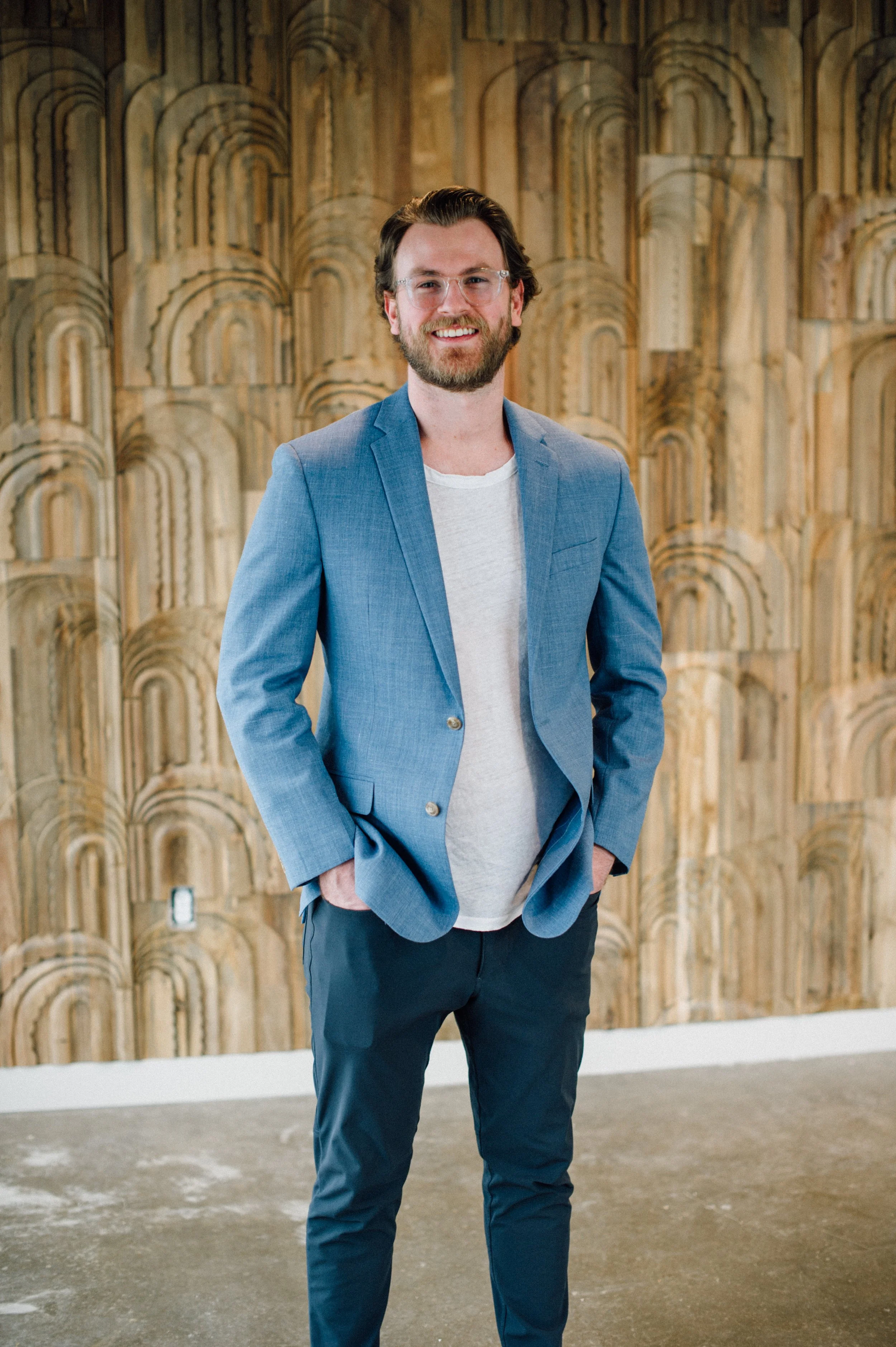 A man with brown hair, beard, and glasses standing in front of a wooden decorative wall, wearing a light blue blazer, white t-shirt, and dark pants, smiling with hands in pockets.
