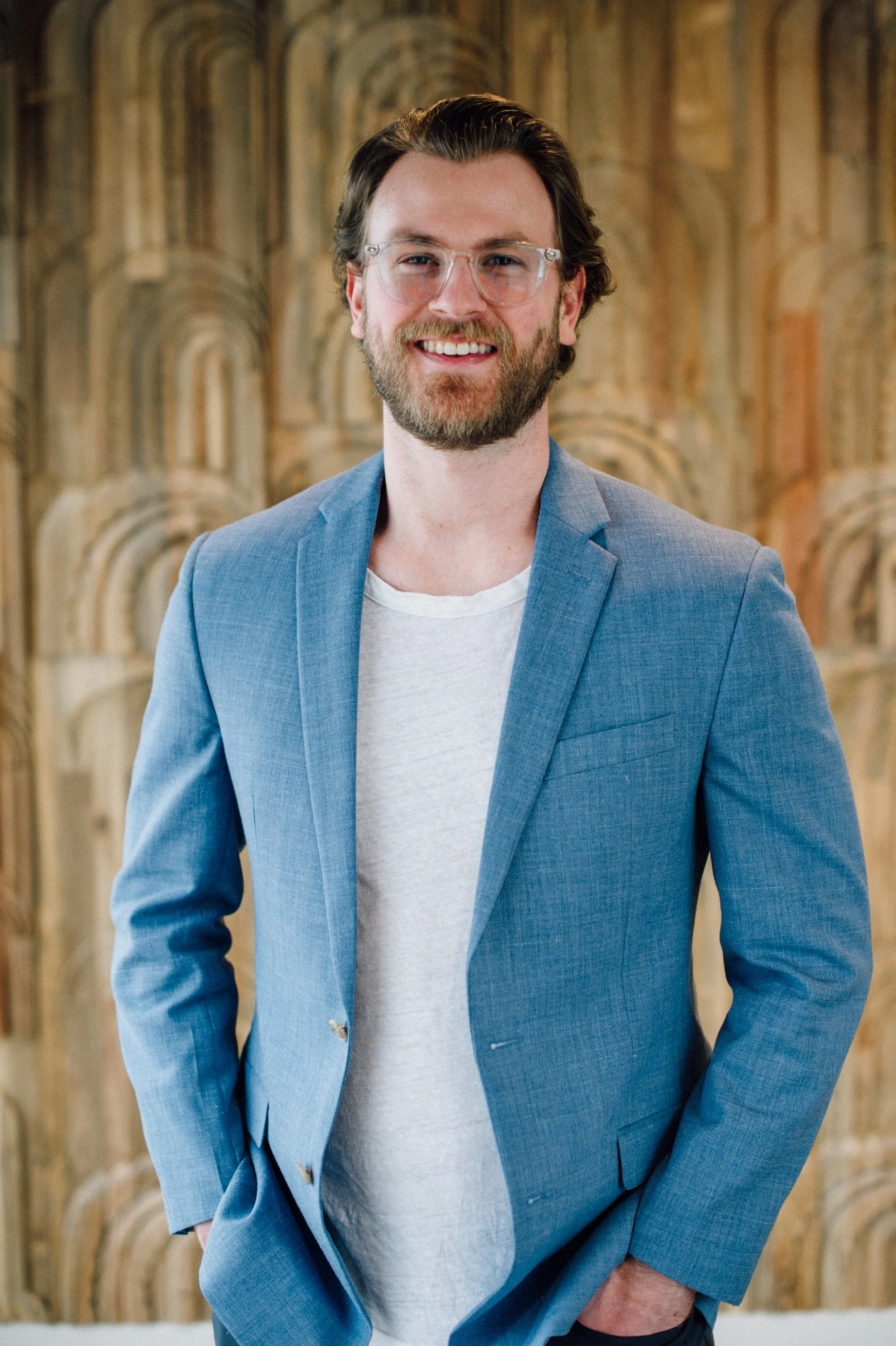 A man with glasses, a beard, and brown hair, wearing a light blue blazer over a white T-shirt, smiling with hands in pockets, standing in front of a decorative wooden wall.