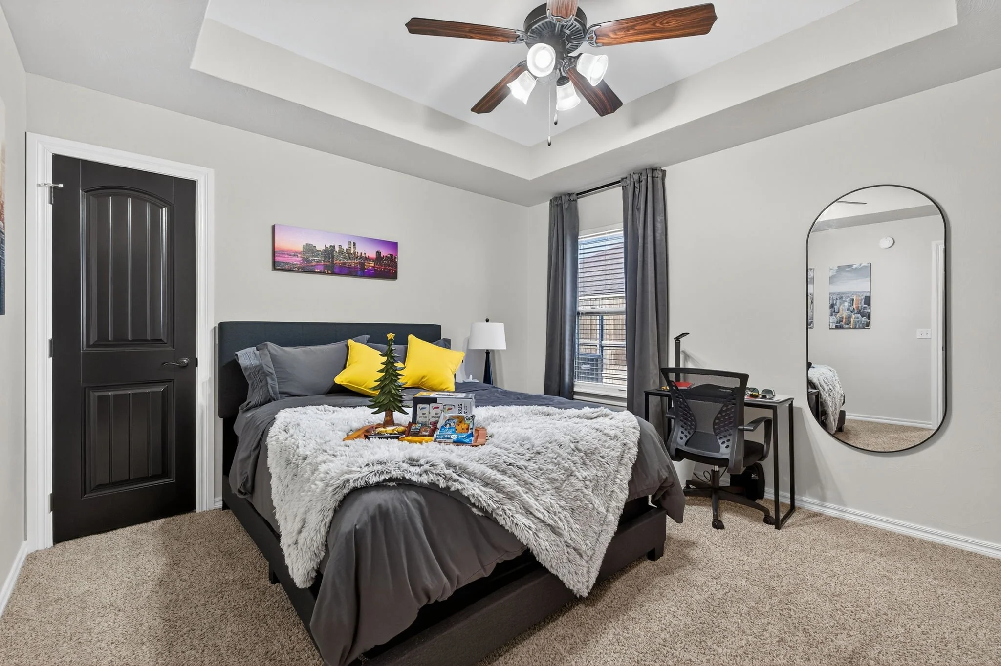 A quiet and peaceful, bedroom with extra pillows, and a plush gray blanket. A window with blinds and magnetic close curtains, a desk with a chair, a lamp, and a large oval mirror on the wall. The ceiling has a fan with wooden blades and lights.
