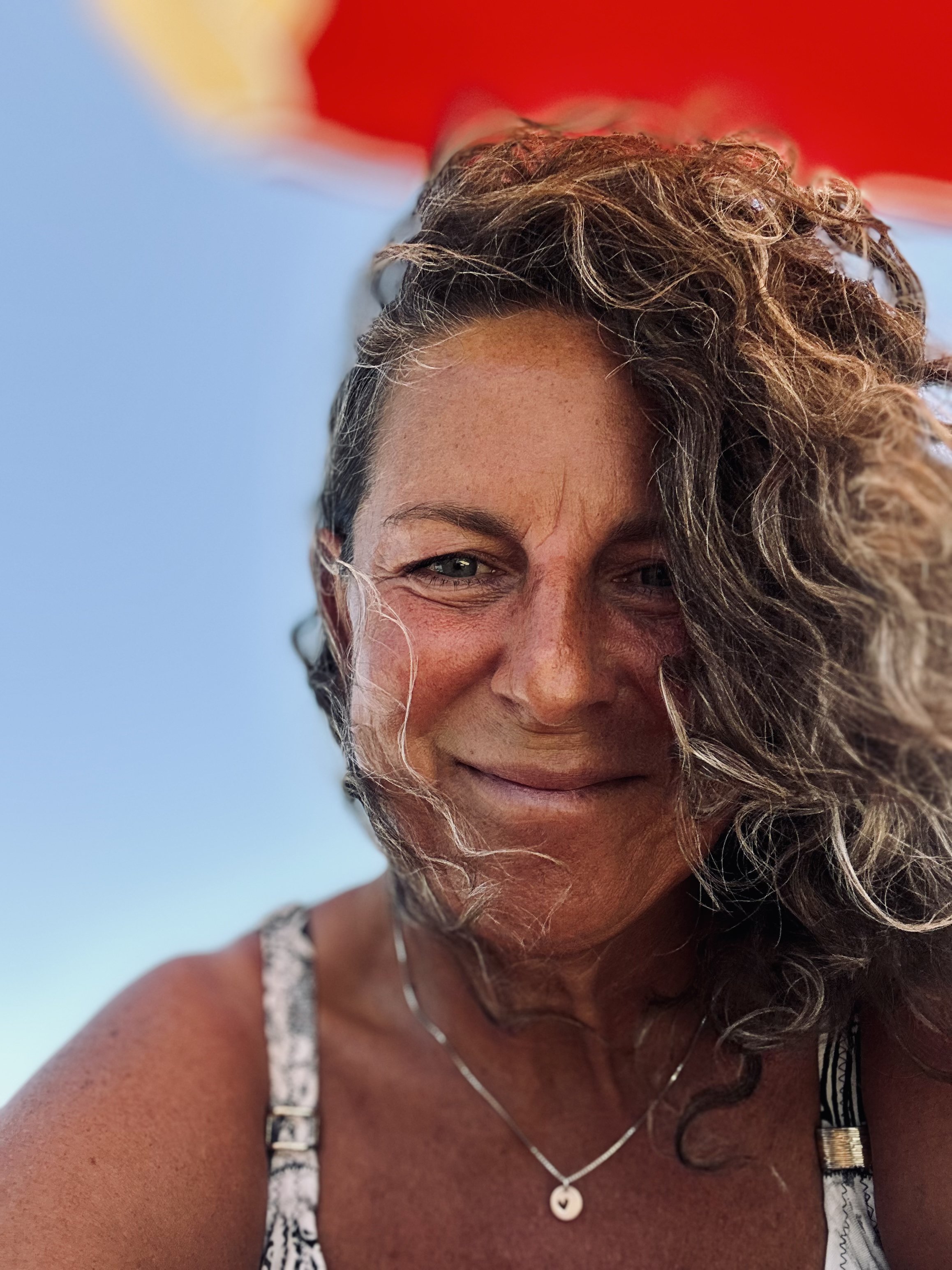 Close-up of a woman with curly hair smiling outdoors, with a red umbrella partially visible at the top and a clear blue sky in the background.