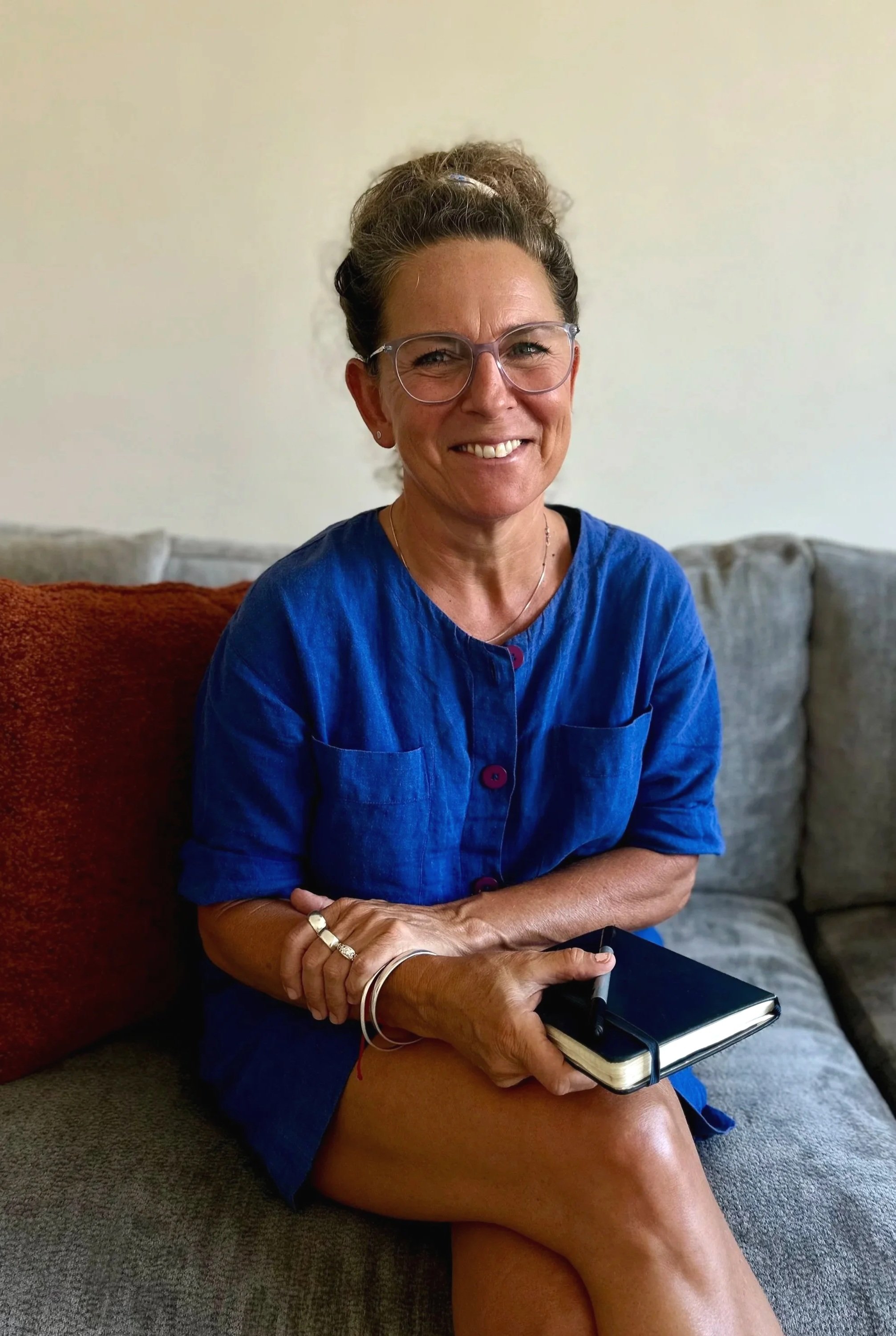 A woman with glasses and a bun hairstyle sitting on a gray couch, smiling, holding a black notebook and pen, wearing a blue button-up shirt.