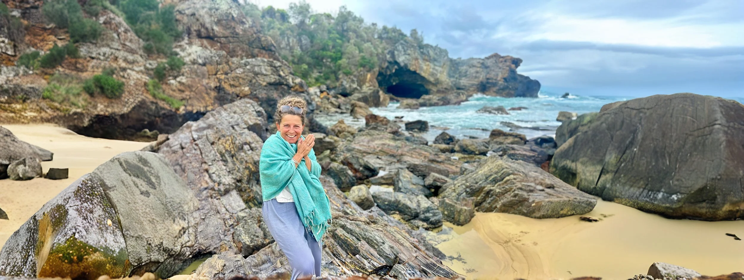 A woman standing on rocks on a beach with a smile, wearing a blue scarf and light-colored pants, with a rocky cliff and ocean waves in the background.