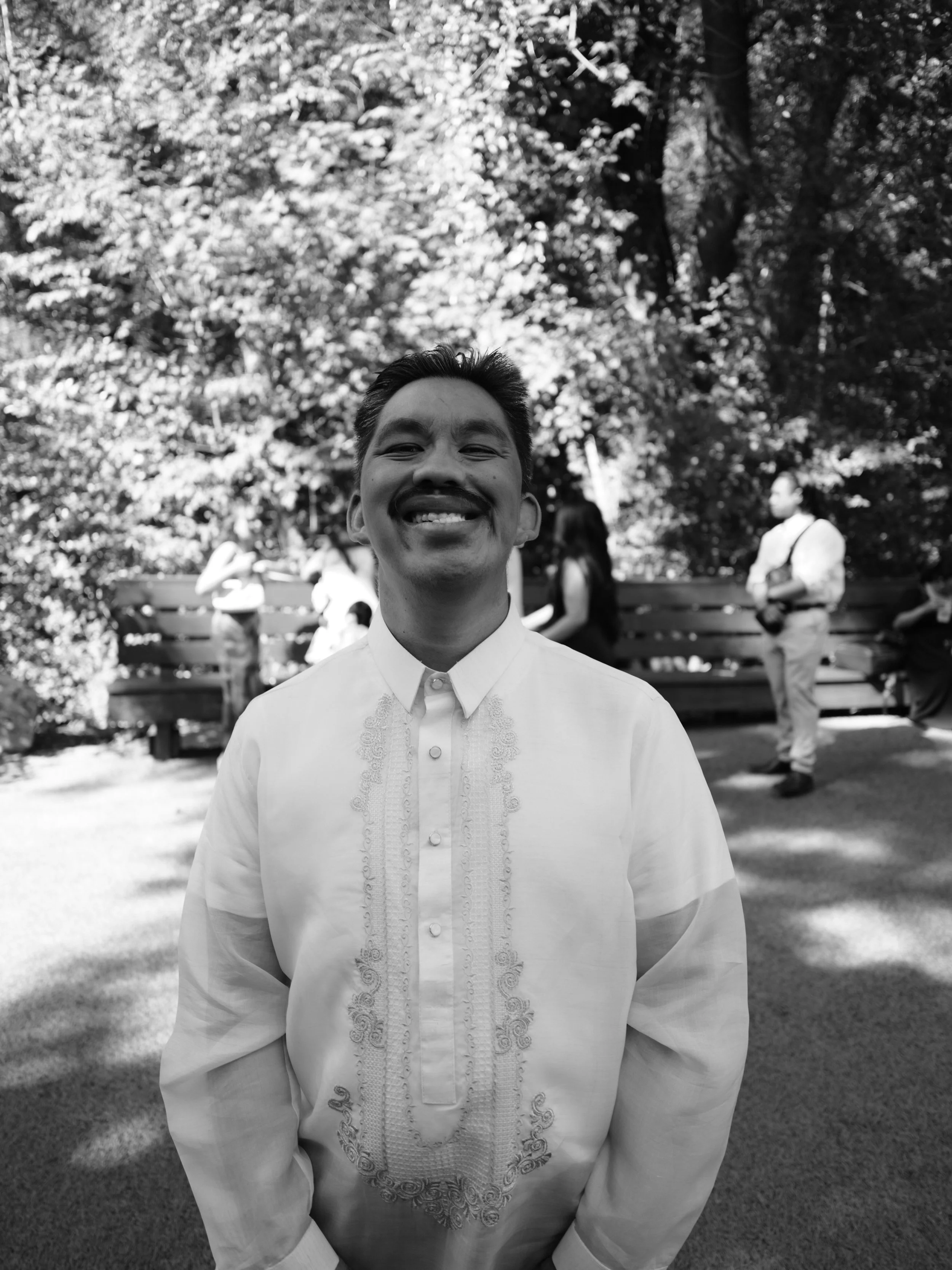 A man smiling in traditional Filipino barong shirt in a park with trees and benches, other people in the background.