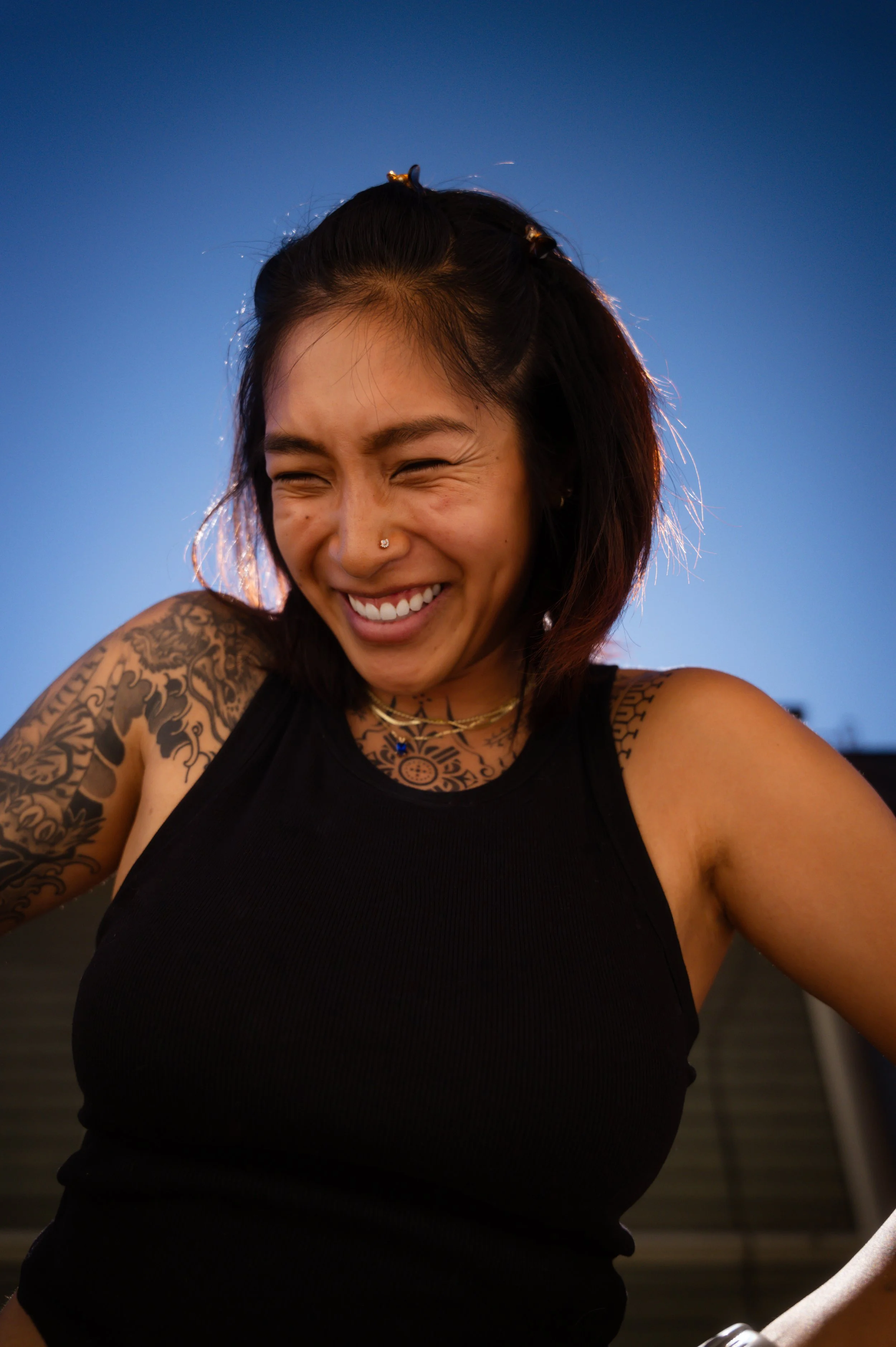 A smiling woman with tattoos on her arm and chest, wearing a black sleeveless top and jewelry, standing outdoors during dusk.
