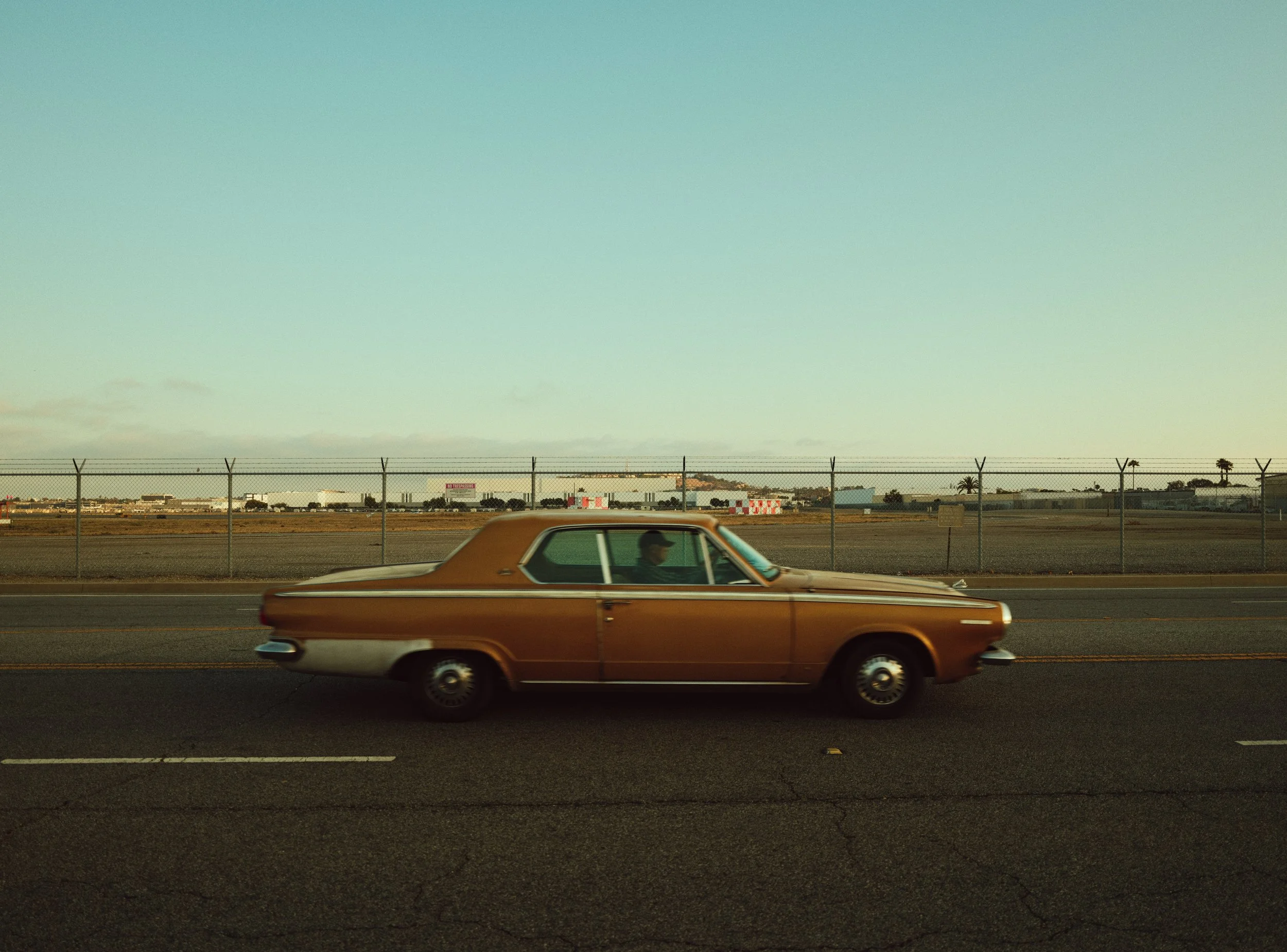 A vintage brown car driving on a road near an airport or industrial area, with a clear sky in the background.
